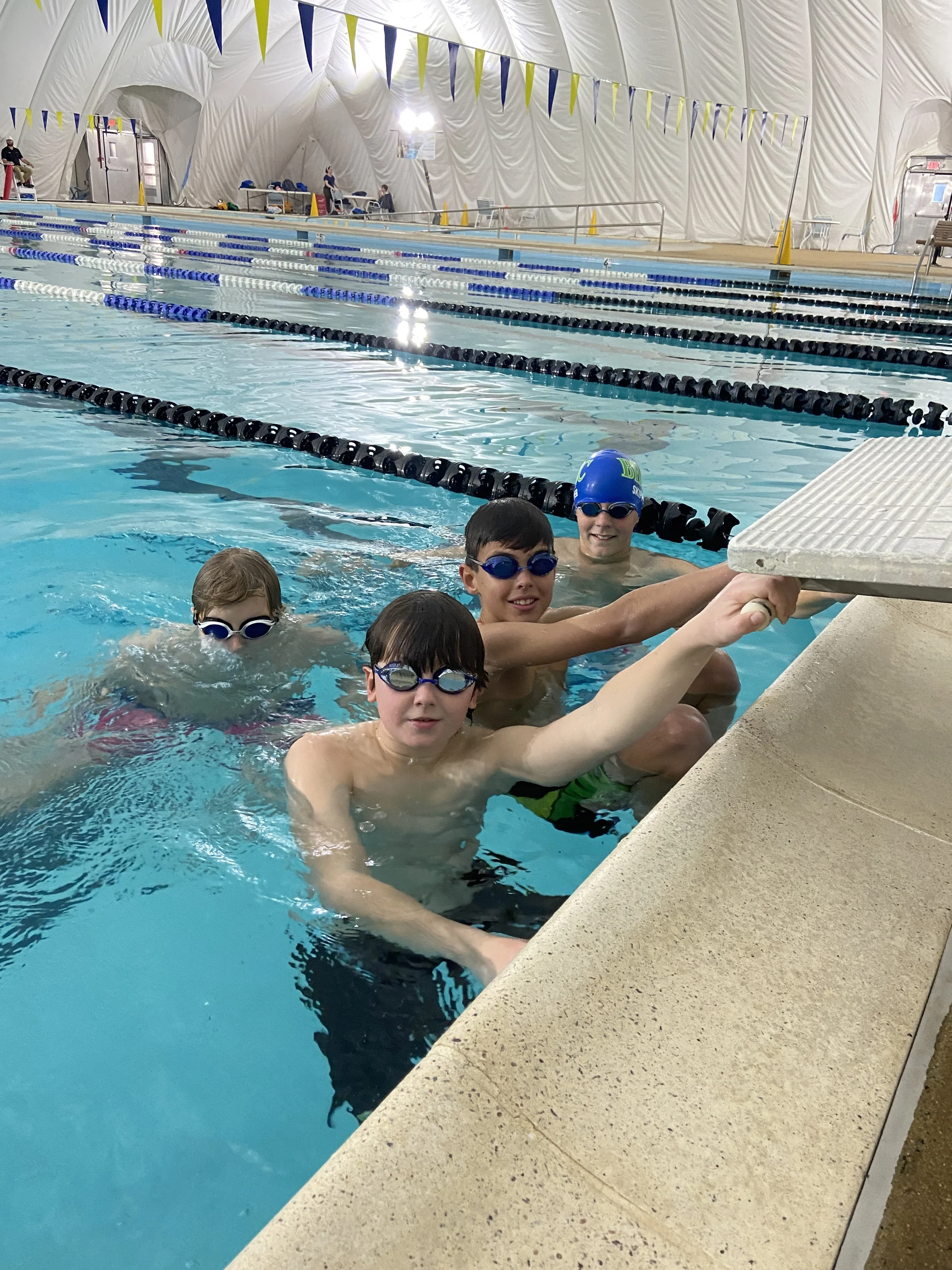 Four children in swimming goggles sitting by the edge of an indoor swimming pool, holding onto the poolside, with a diving board nearby.