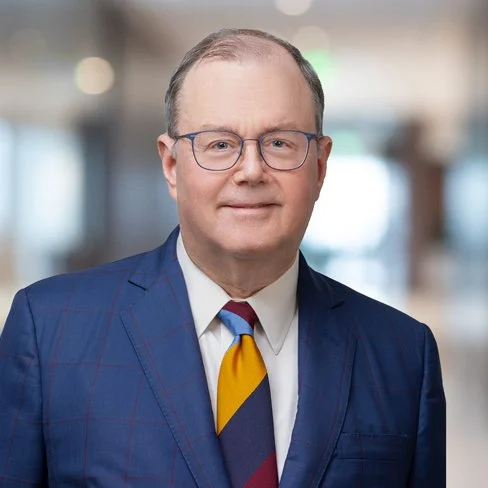 Portrait of a middle-aged man wearing glasses, a dark blue suit, a white shirt, and a colorful tie, standing in a blurred indoor background.