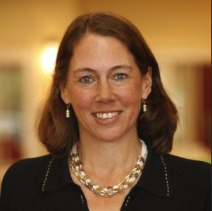 A woman with shoulder-length brown hair, wearing earrings, a pearl necklace, and a dark blazer, smiling indoors with a blurred background.