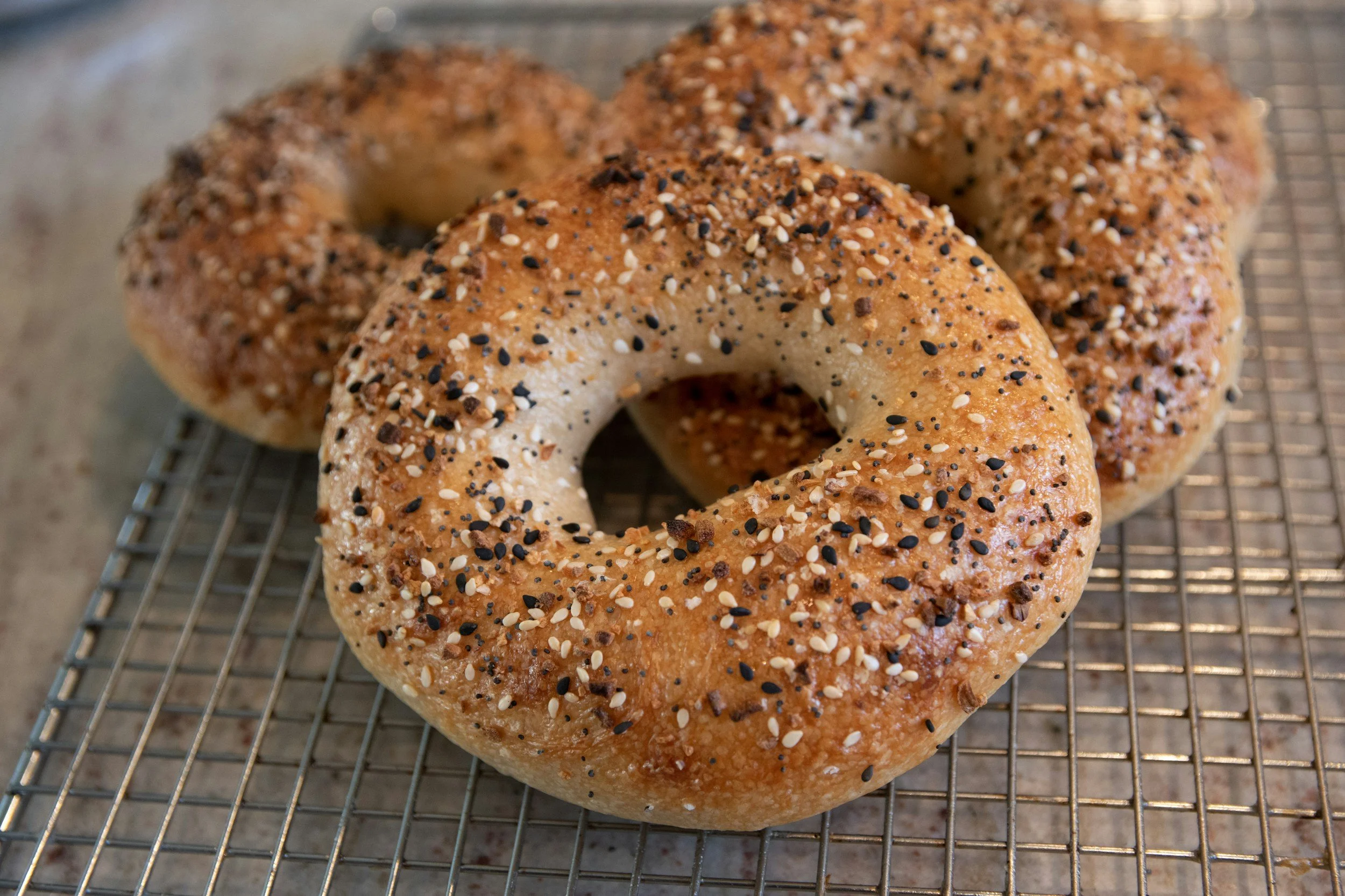 Three freshly baked bagels sprinkled with various seeds and spices, resting on a wire cooling rack.