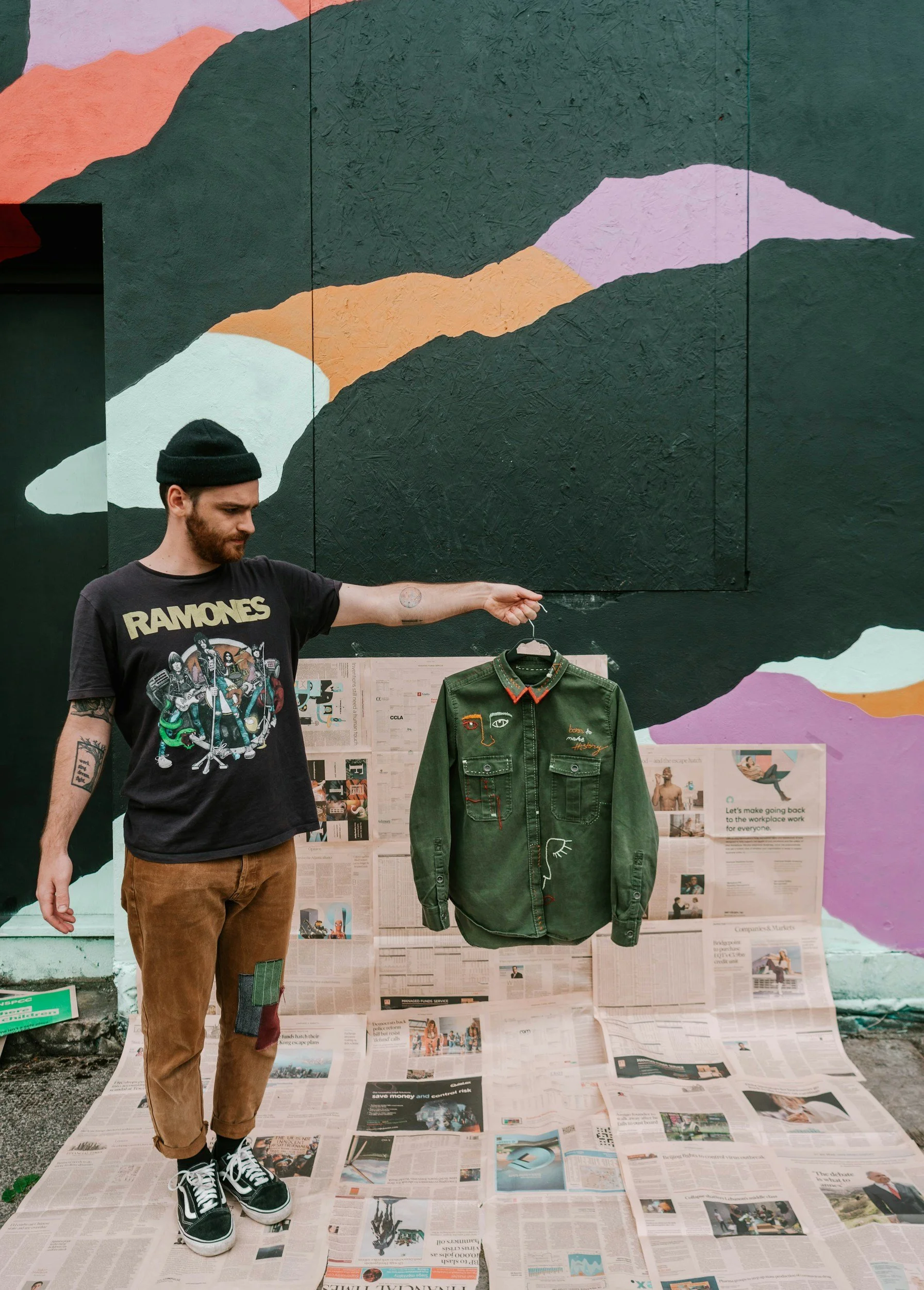 A man with a beard wearing a black beanie, Ramones t-shirt, and brown pants, is holding a green denim jacket up with a hanger. The jacket has embroidered designs including a face, eyes, a heart, and a hand. The background features a colorful mural on a wall, and newspapers are spread out on the ground behind him.