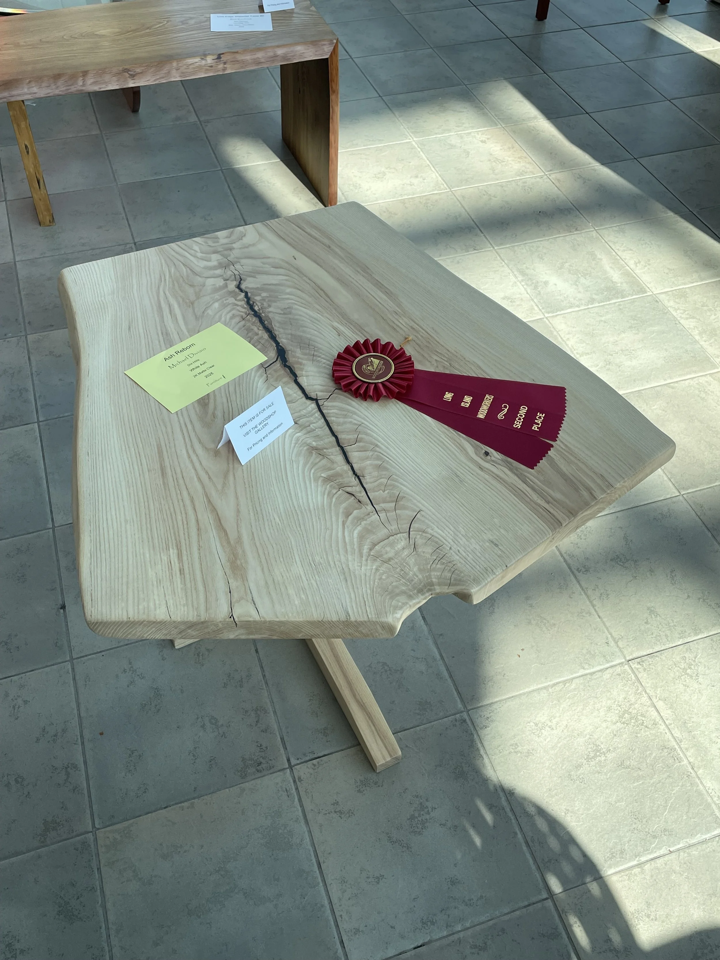 A wooden table with a prominent crack running down the center, displaying a red ribbon rosette with a gold emblem. The table is in a well-lit room with tiled flooring, and there are labels on the table indicating it is an exhibit for an art or woodwo