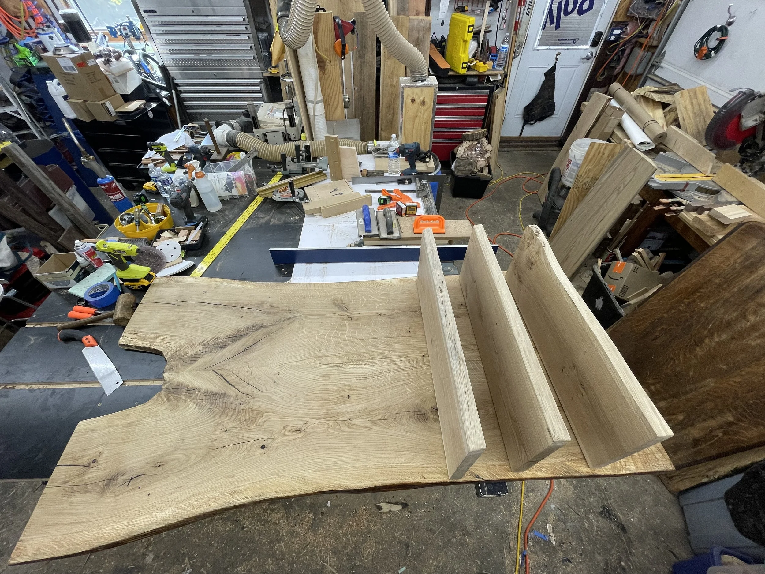 A woodworking workshop with a large, irregularly shaped wooden slab and three smaller wooden pieces on a workbench. The cluttered workspace contains various tools, measuring tapes, clamps, and supplies scattered across the table, with shelves, storag