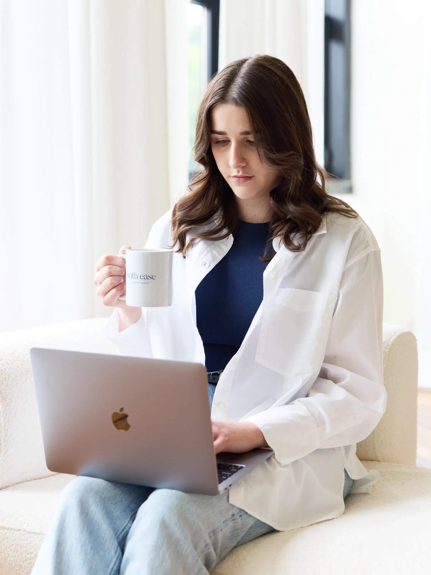 Taylor of With Ease Therapy Space in Guelph holding a mug and on a MacBook in a comfy chair.