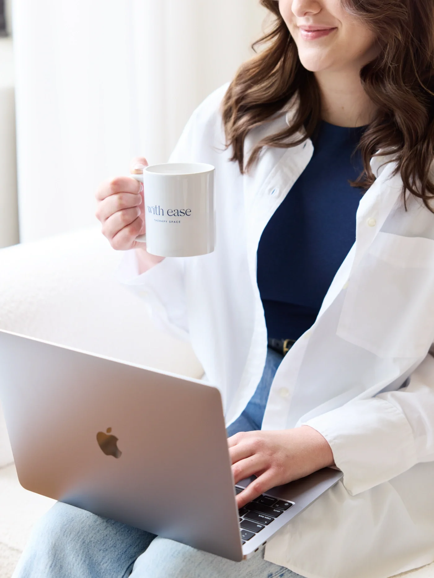 Taylor of With Ease Therapy Space in Guelph holding a mug and on a MacBook in a comfy chair.
