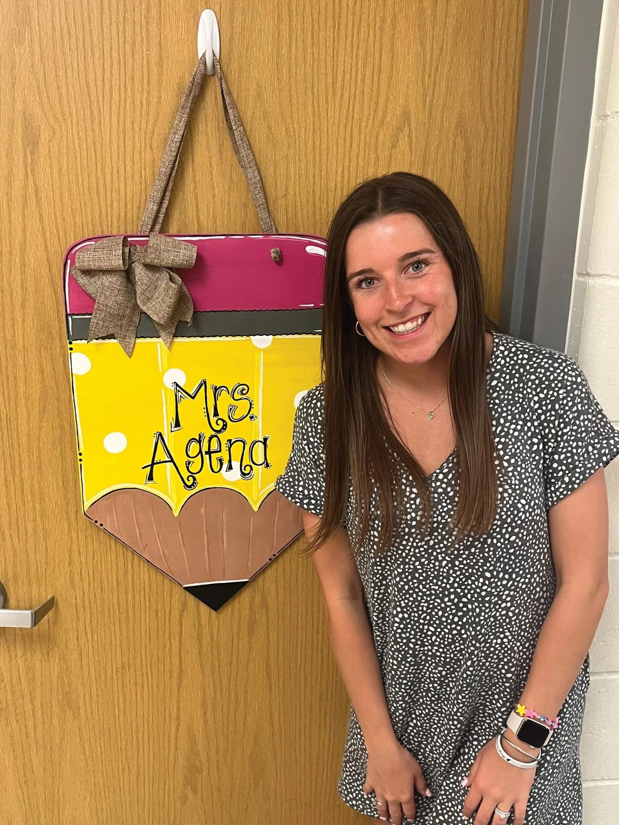 Mrs. Agena standing in front of her classroom door which has a personalized pencil sign with her name on it