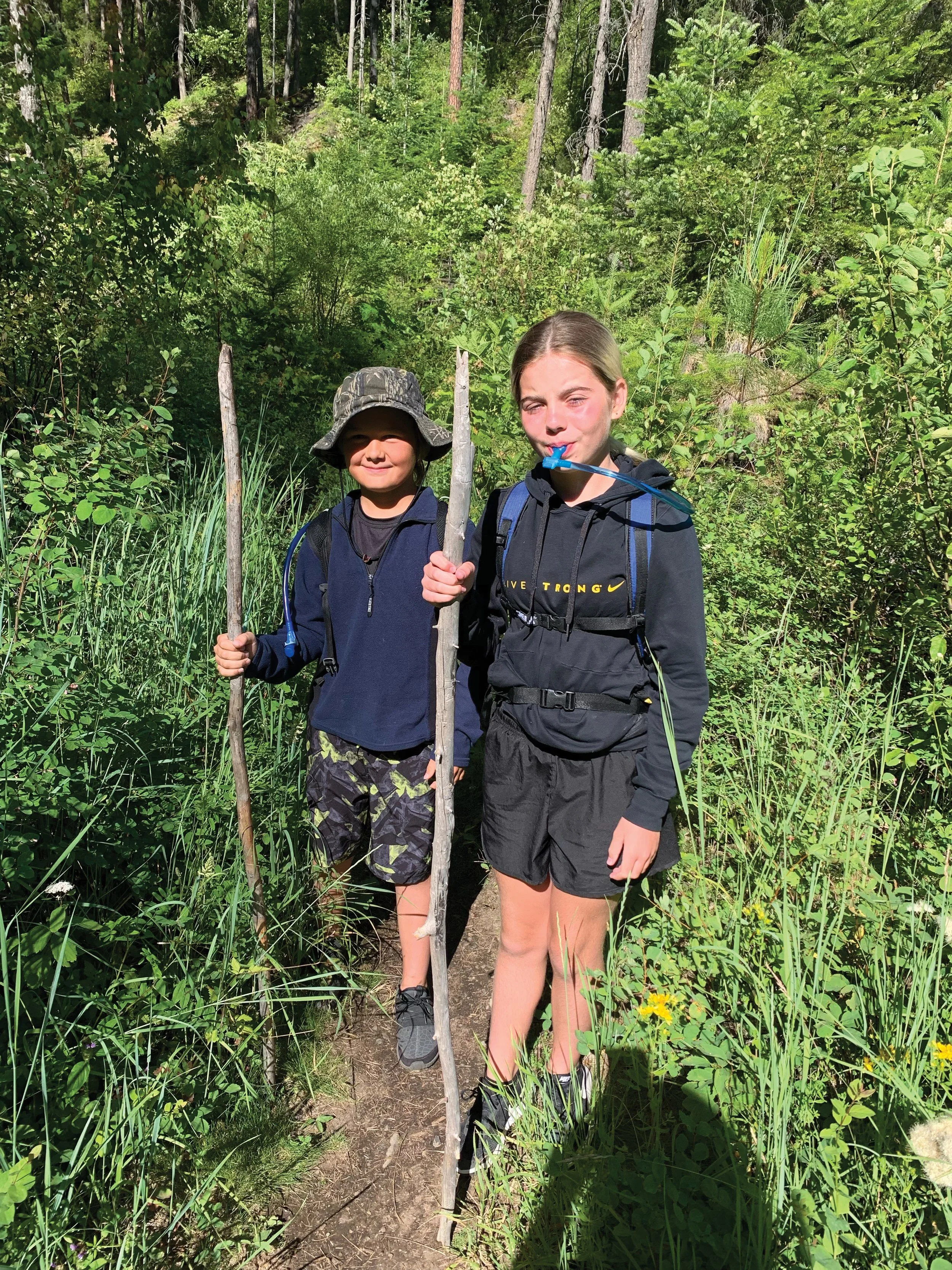 two kids (one boy, one girl) standing with wooden walking sticks in the woods