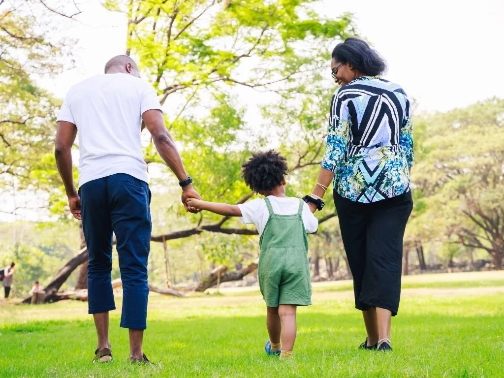 Two parents handing hands with their young child outside in a park