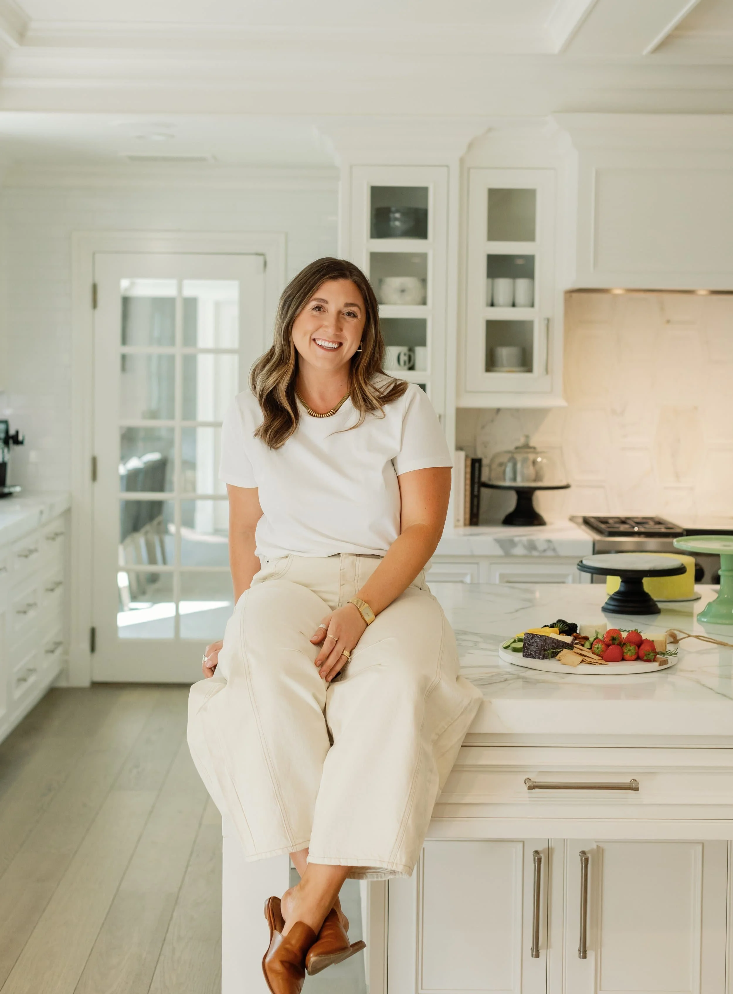 Dietician in CA offering virtual nutrition counseling sitting on a kitchen counter, smiling, wearing a white t-shirt and cream pants, with a charcuterie plate and strawberries nearby.