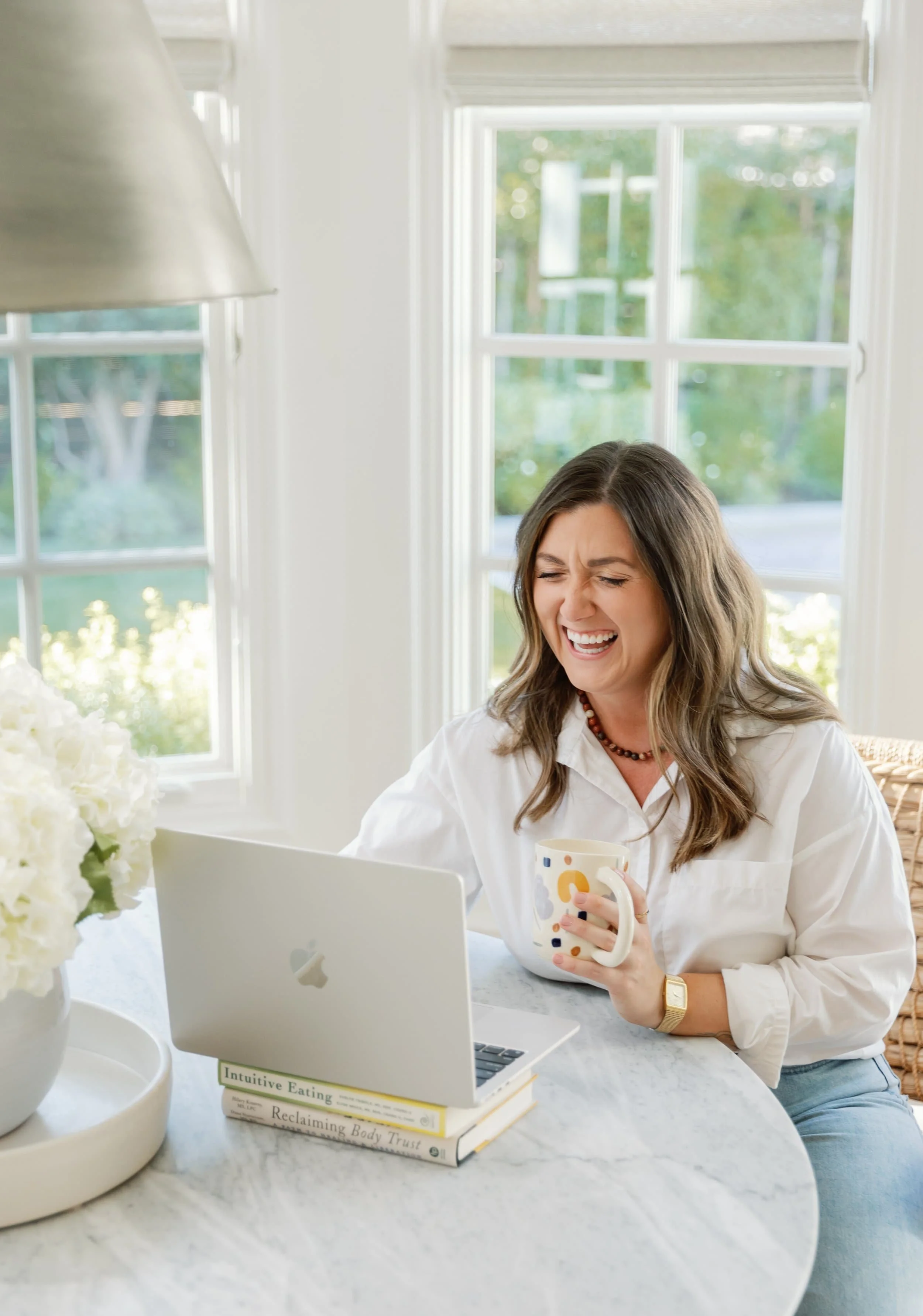 Dietician nutritionist in CA laughing while sitting at a kitchen table with a laptop and holding a mug, with large windows in the background.