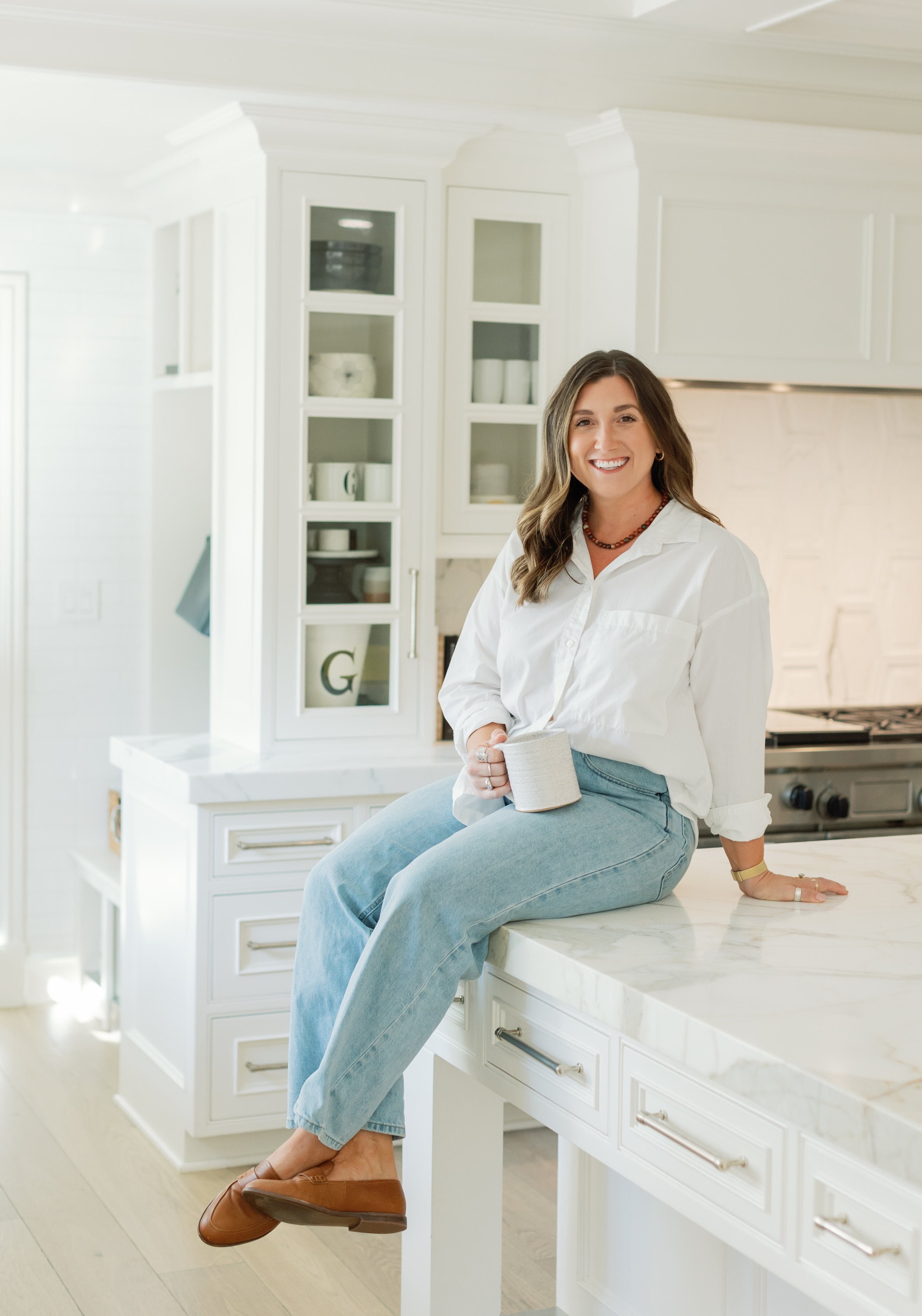 Dietician nutritionist specializing in anorexia with wavy brown hair smiling while sitting on a kitchen countertop, holding a mug, wearing a white button-up shirt, light blue jeans, and brown flats in a bright, modern kitchen.