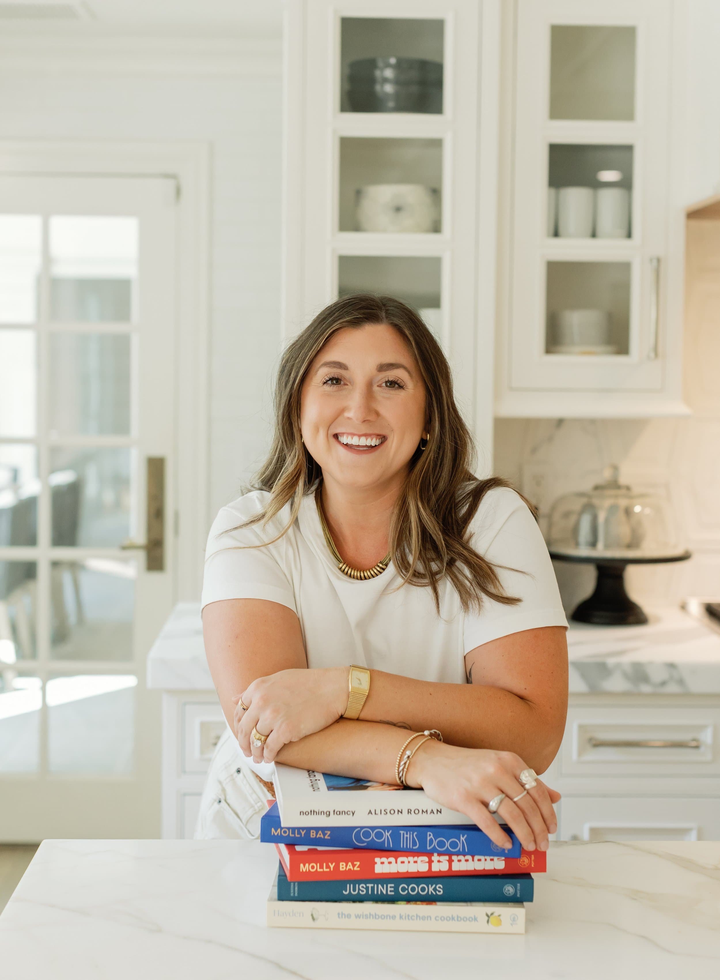 Dietitian Nutritionist who specializes in eating disorders smiling in a kitchen resting her arms on a stack of five cookbooks on a white marble island.