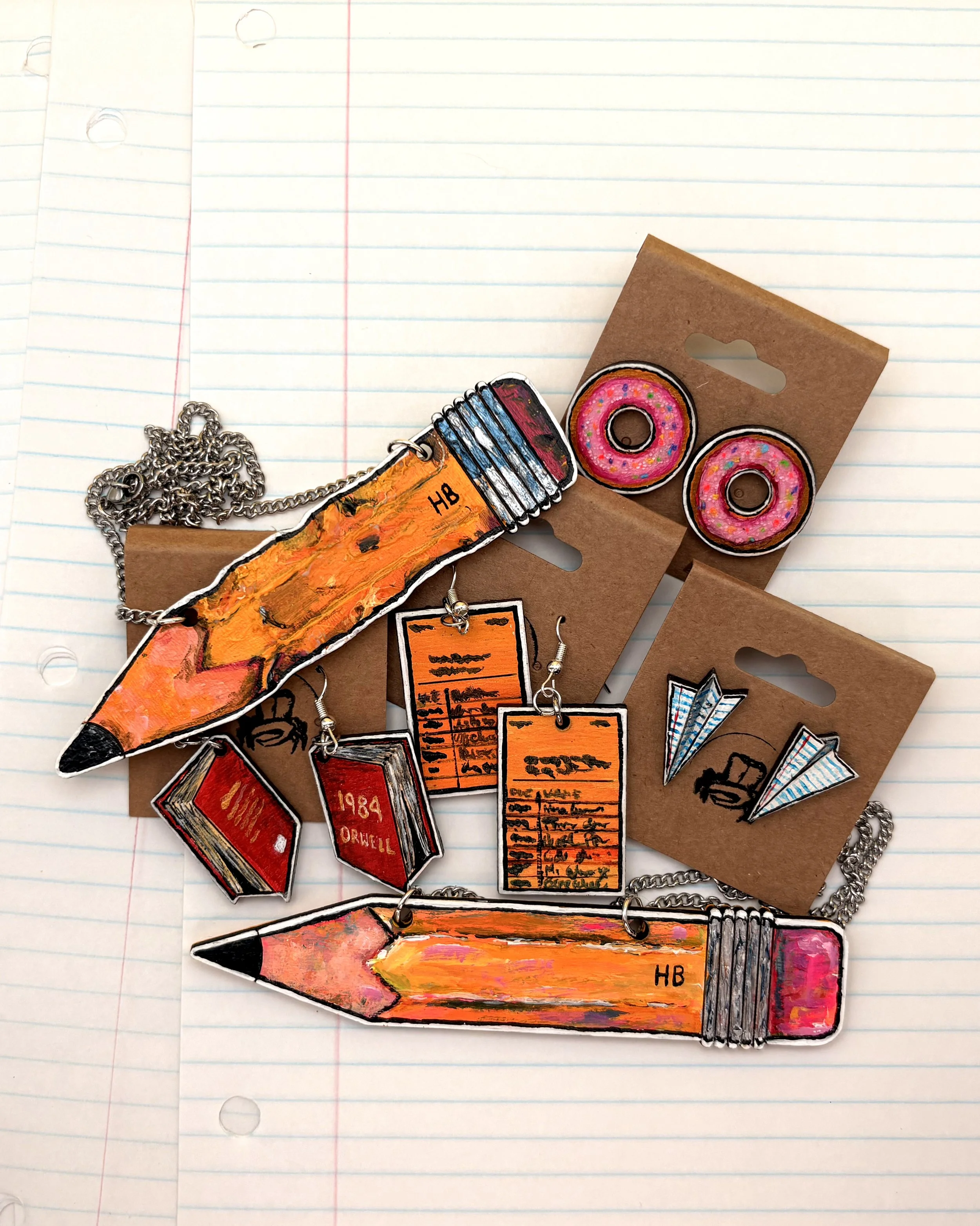 Colorful jewelry charms in the shape of a pencil, a donut, and a license plate, placed on lined notebook paper.