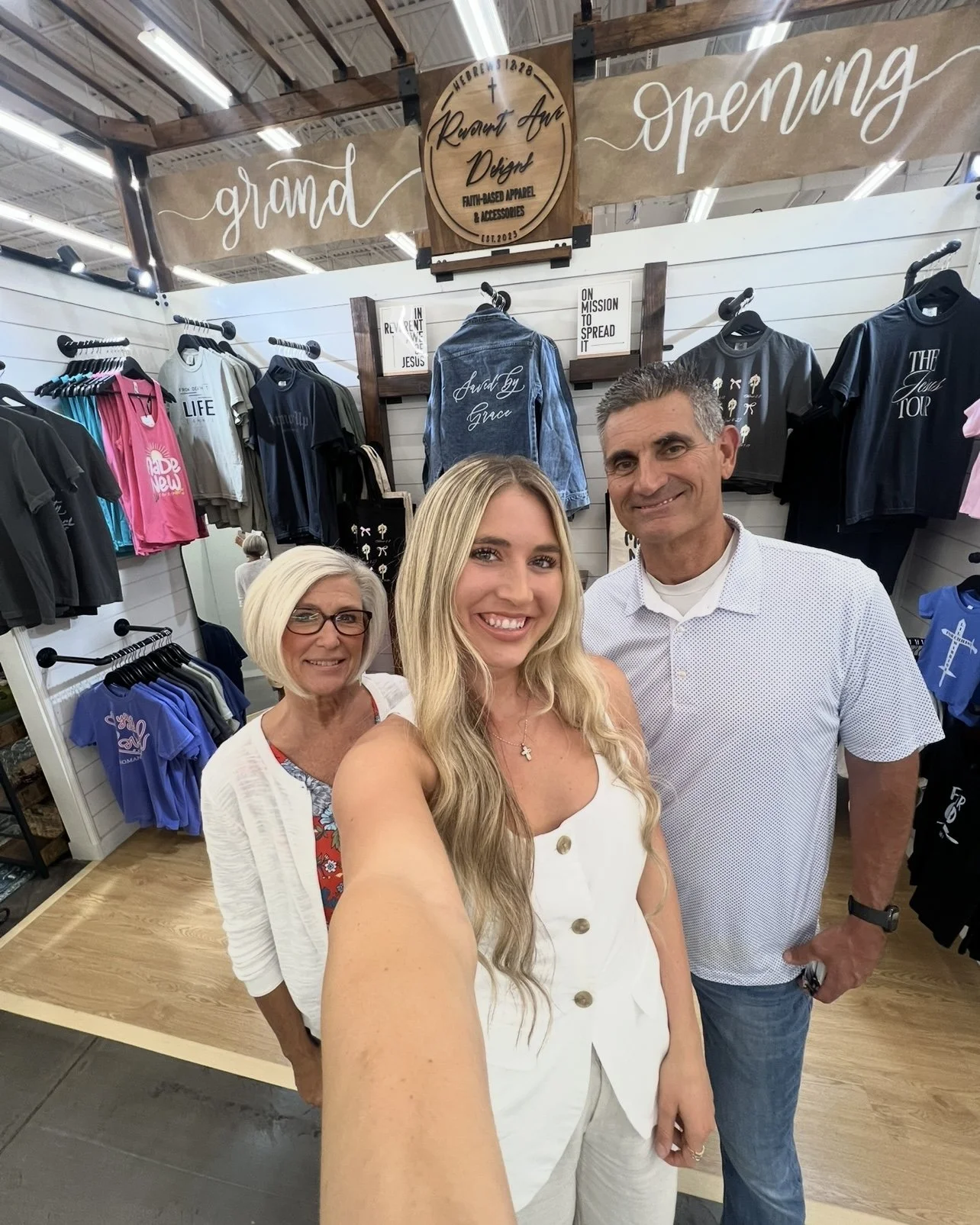 Three people smiling for a selfie inside a clothing store with various T-shirts and jackets displayed on the wall behind them.