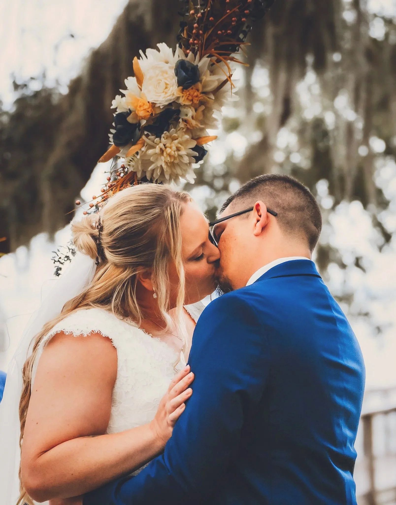 A couple is kissing outdoors during their wedding, with the bride’s blonde hair in a loose braid and wearing a white lace dress, and the groom wearing glasses and a blue suit. They are holding hands, and a floral garland is hanging above them, with t