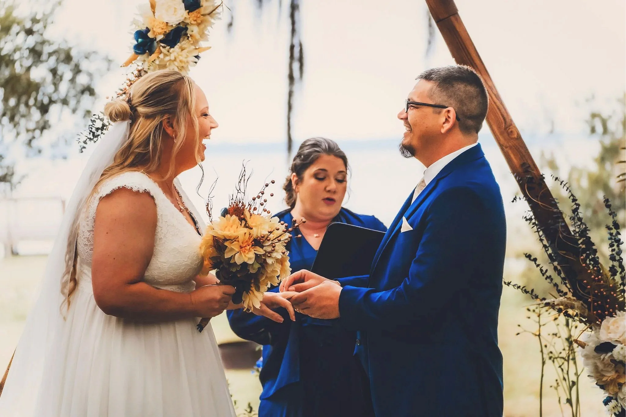 A couple getting married outdoors, exchanging vows, with a woman officiating, a wooden arch decorated with flowers in the background, and the bride holding a bouquet. The bride has long blonde hair and is wearing a white wedding dress, and the groom 