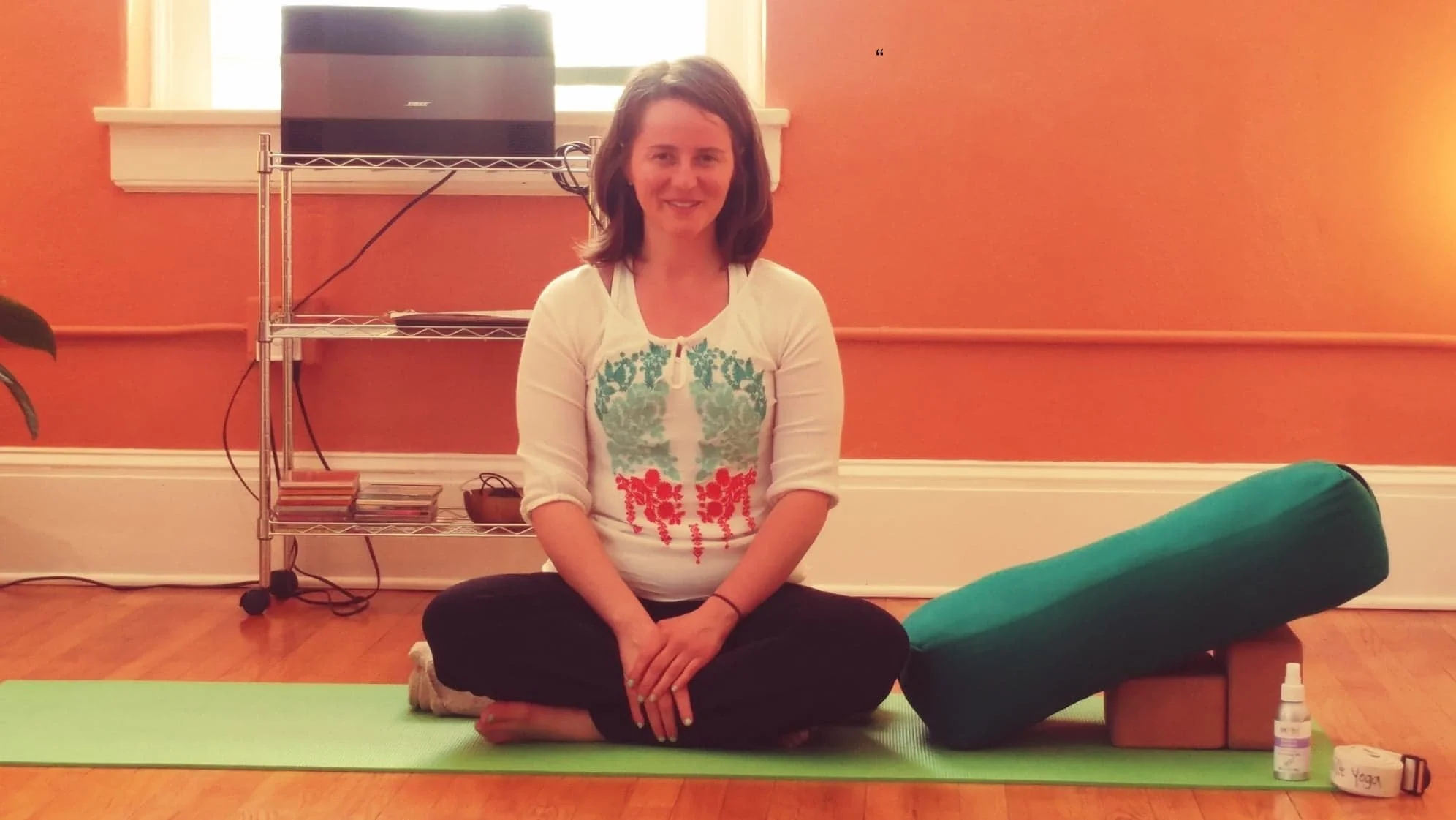Nina Marie Collins sitting cross-legged on a yoga mat in a room, with a bolster and spray bottle nearby, smiling at the camera.