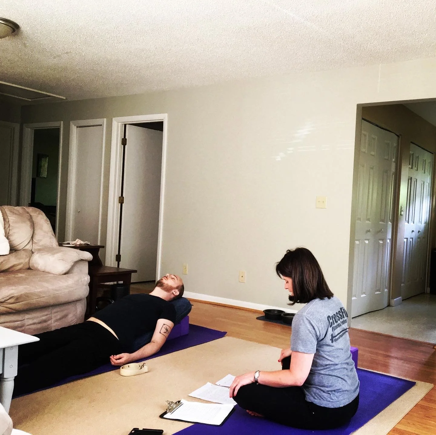 A woman sitting on a yoga mat with a clipboard and papers in front of her, observing a man lying on a yoga mat with his eyes closed, inside a living room.