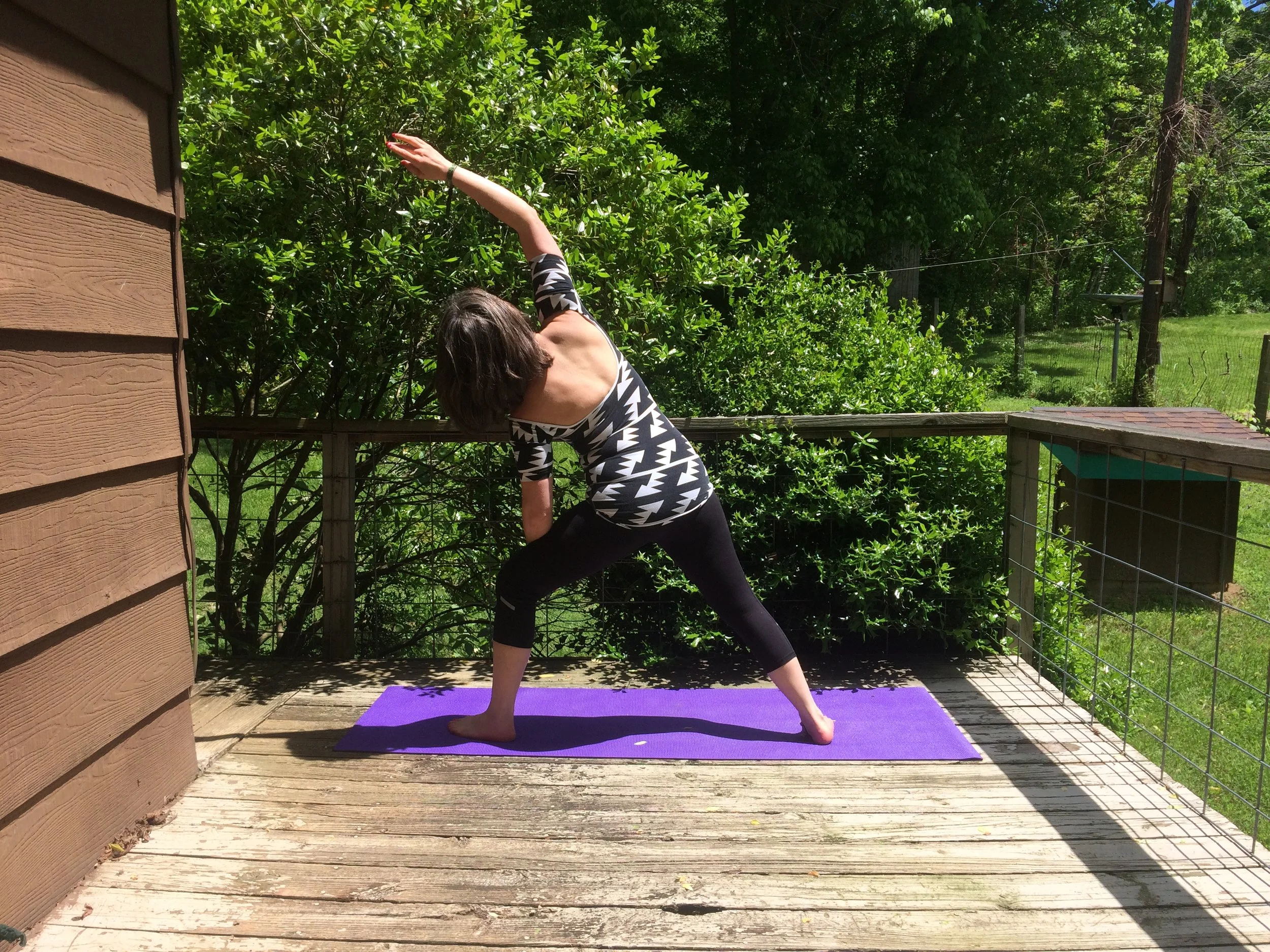 Nina Marie Collins practicing yoga on a wooden deck outdoors, performing a side bend stretch on a purple yoga mat, with lush green trees and shrubbery in the background.