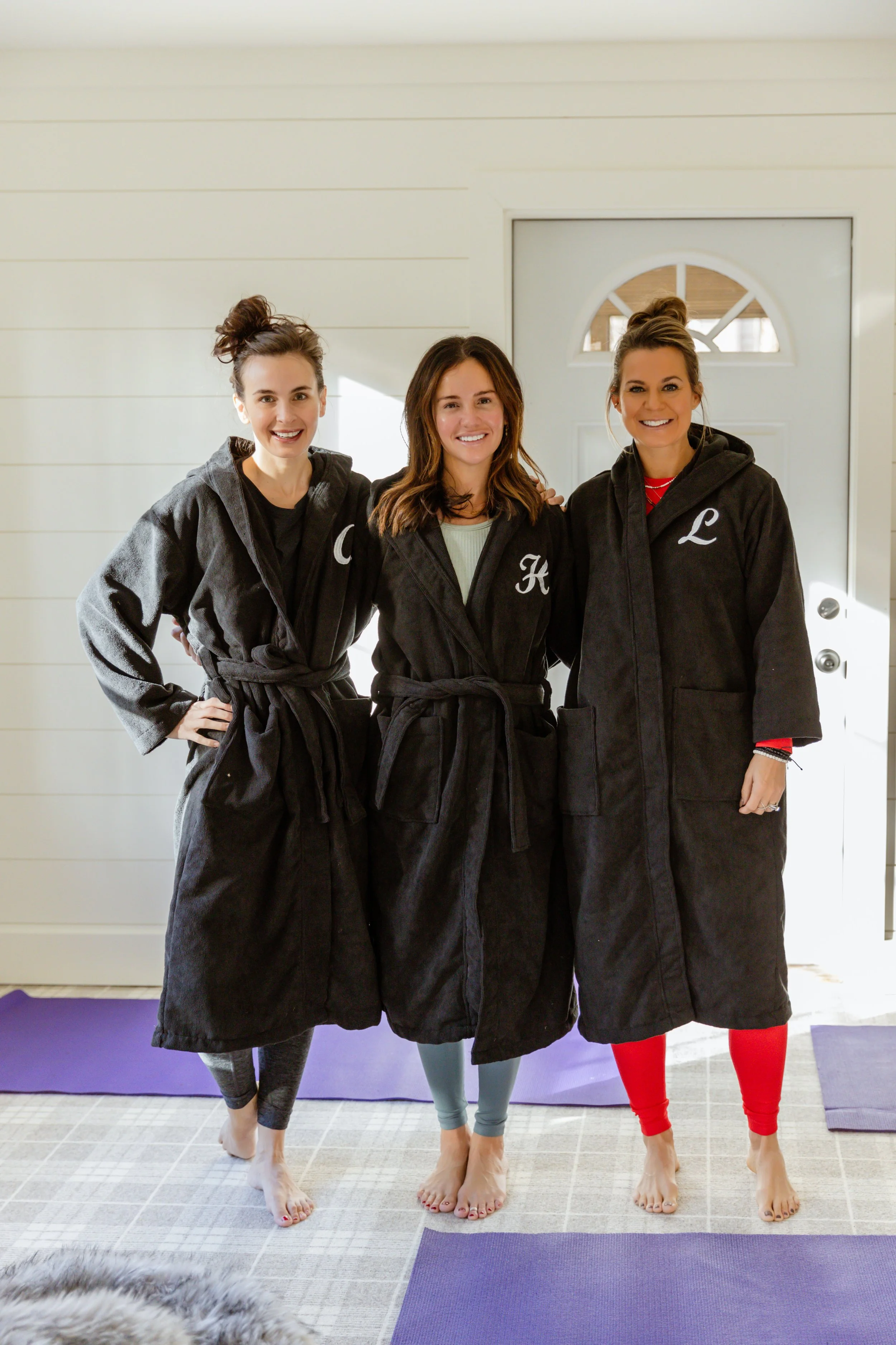 Three women standing indoors in front of a door, wearing black bathrobes with initials, smiling, and barefoot on yoga mats.