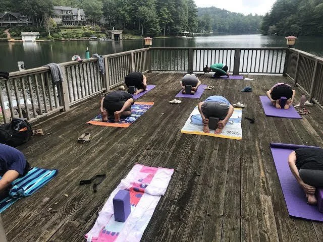 People practicing yoga on a wooden deck by a lake, with trees and houses in the background.