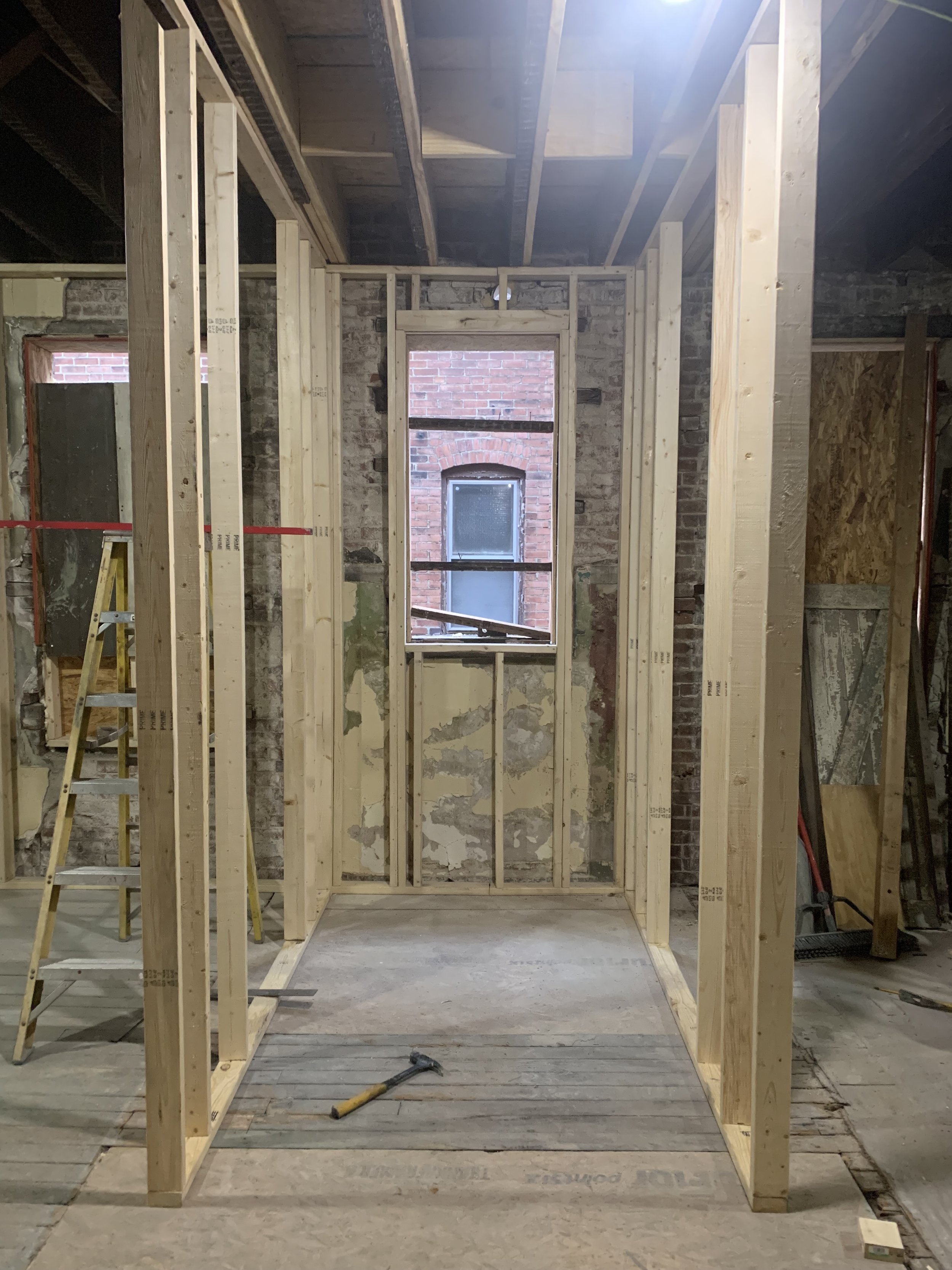 Interior of a building under construction with wooden framing, an unfinished wall, and exposed brick wall visible through a window opening.