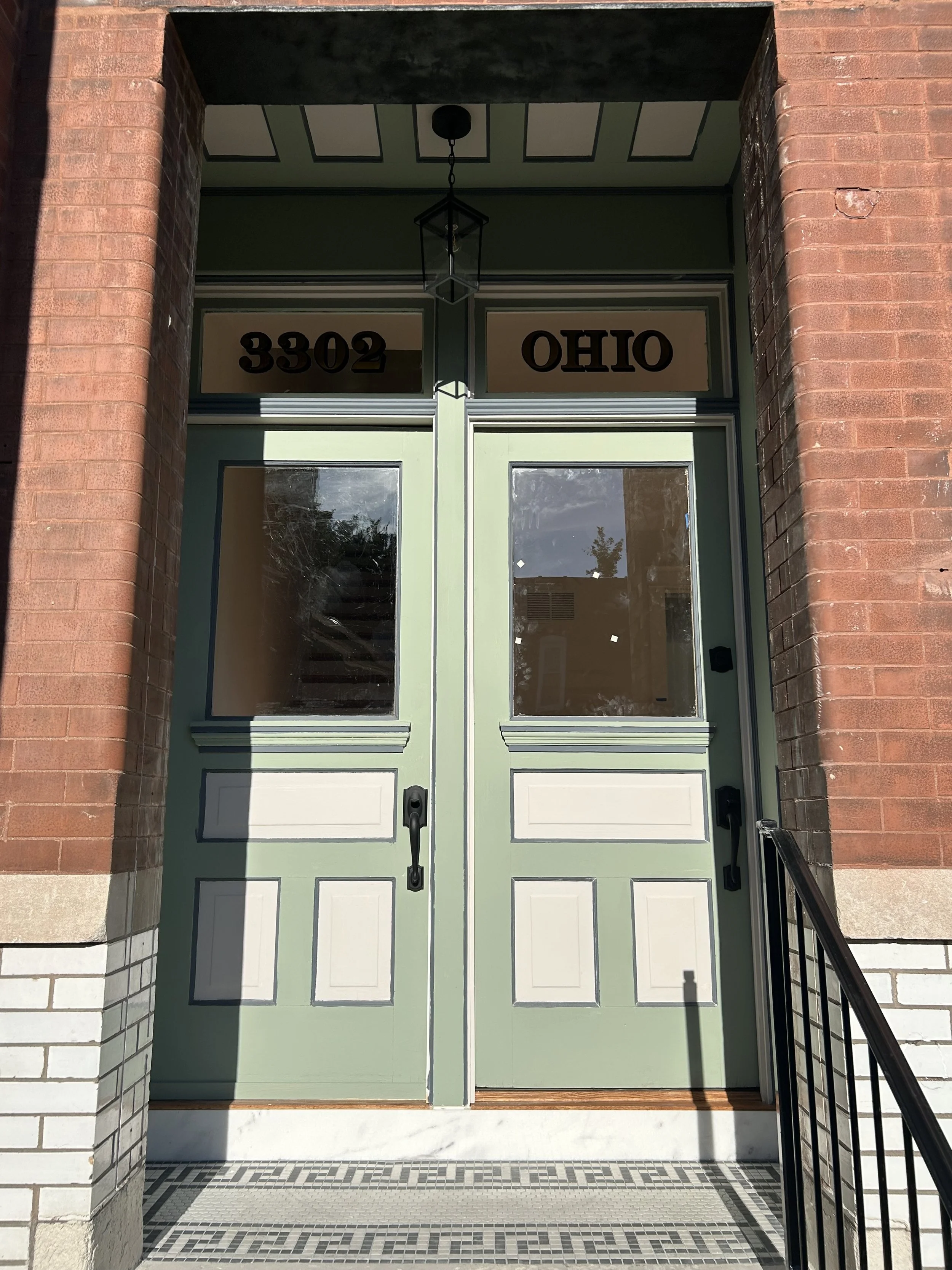 Green double front doors with black handles, surrounded by a brick entryway, with house numbers 3302 and Ohio above the doors, a hanging lantern light fixture overhead, and a tiled concrete porch with steps and black railing on the right.
