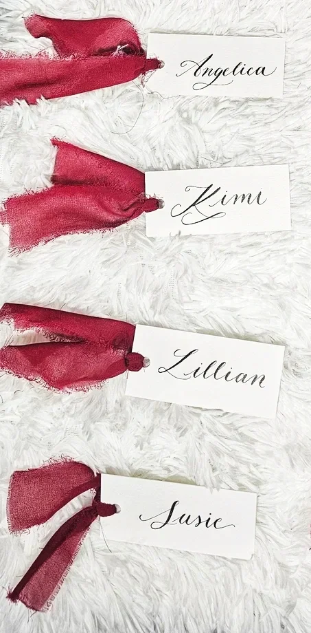 Four white cards with elegant cursive names: Angelica, Kimi, Lillian, Susie, each attached to red fabric ribbons with frayed edges, laid on a white, fluffy surface.