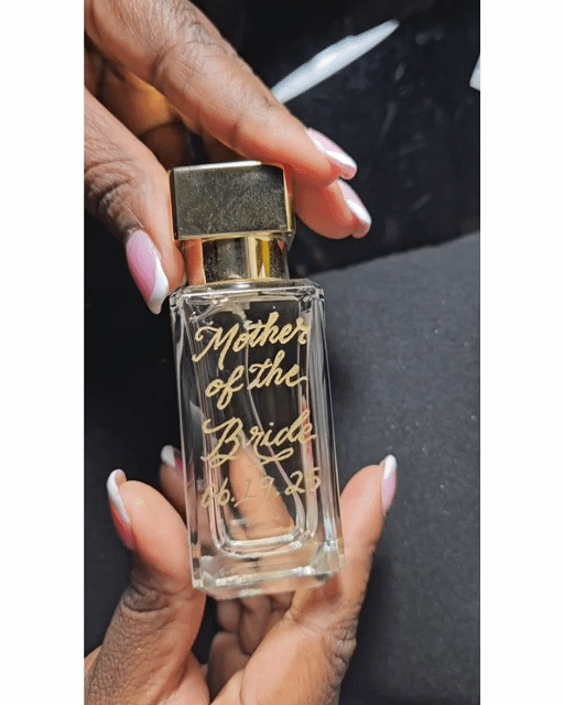 A hand holding a glass perfume bottle with gold lettering that reads "Mother of the Bride" and a date. The person has light nail polish.