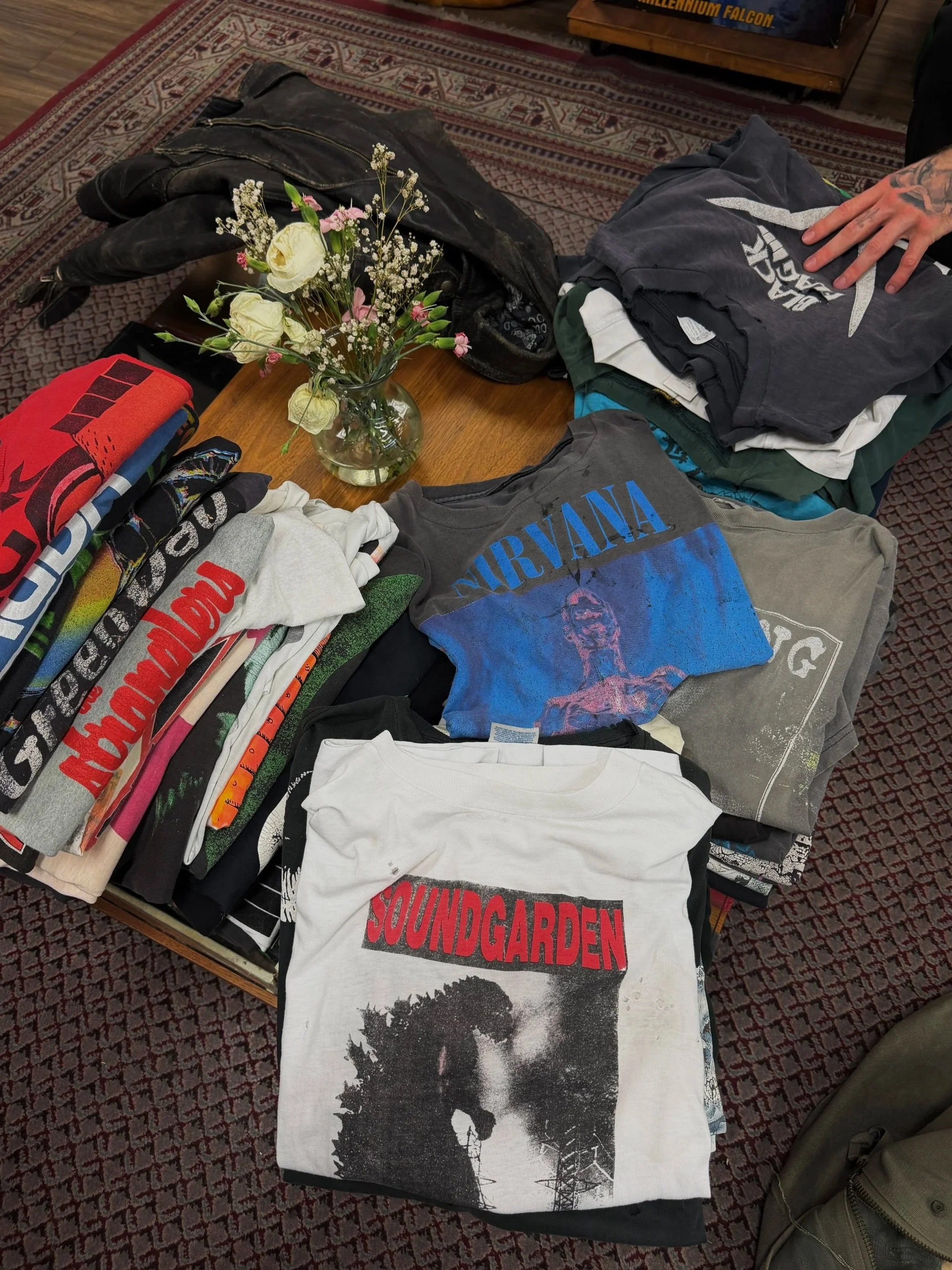 A table with a variety of band T-shirts, folded and spread out, including Nirvana and Soundgarden, alongside a vase of flowers, and some folded clothes and a leather jacket on the table.