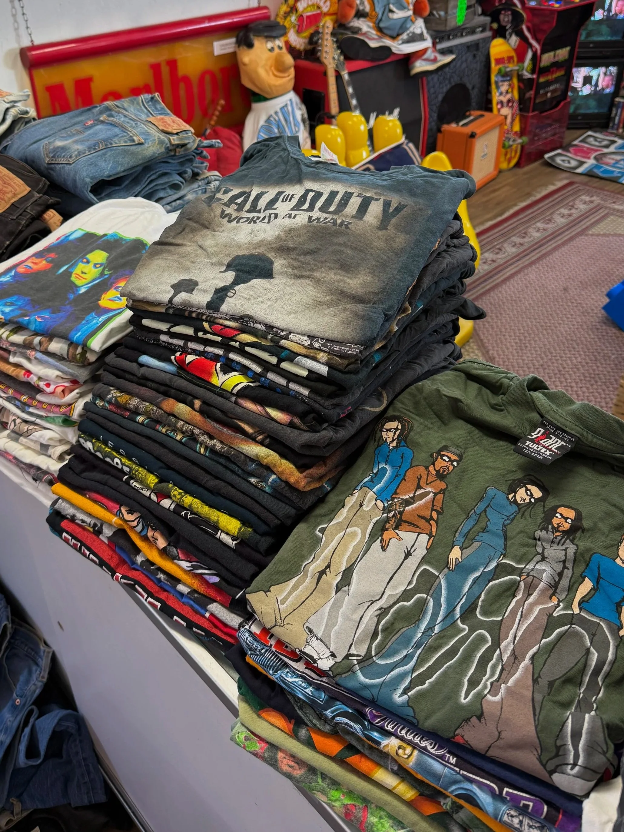 A table filled with folded graphic T-shirts featuring pop culture designs, including a Call of Duty shirt, a rock band print, and characters from animated shows, in a vintage clothing or thrift store setting.