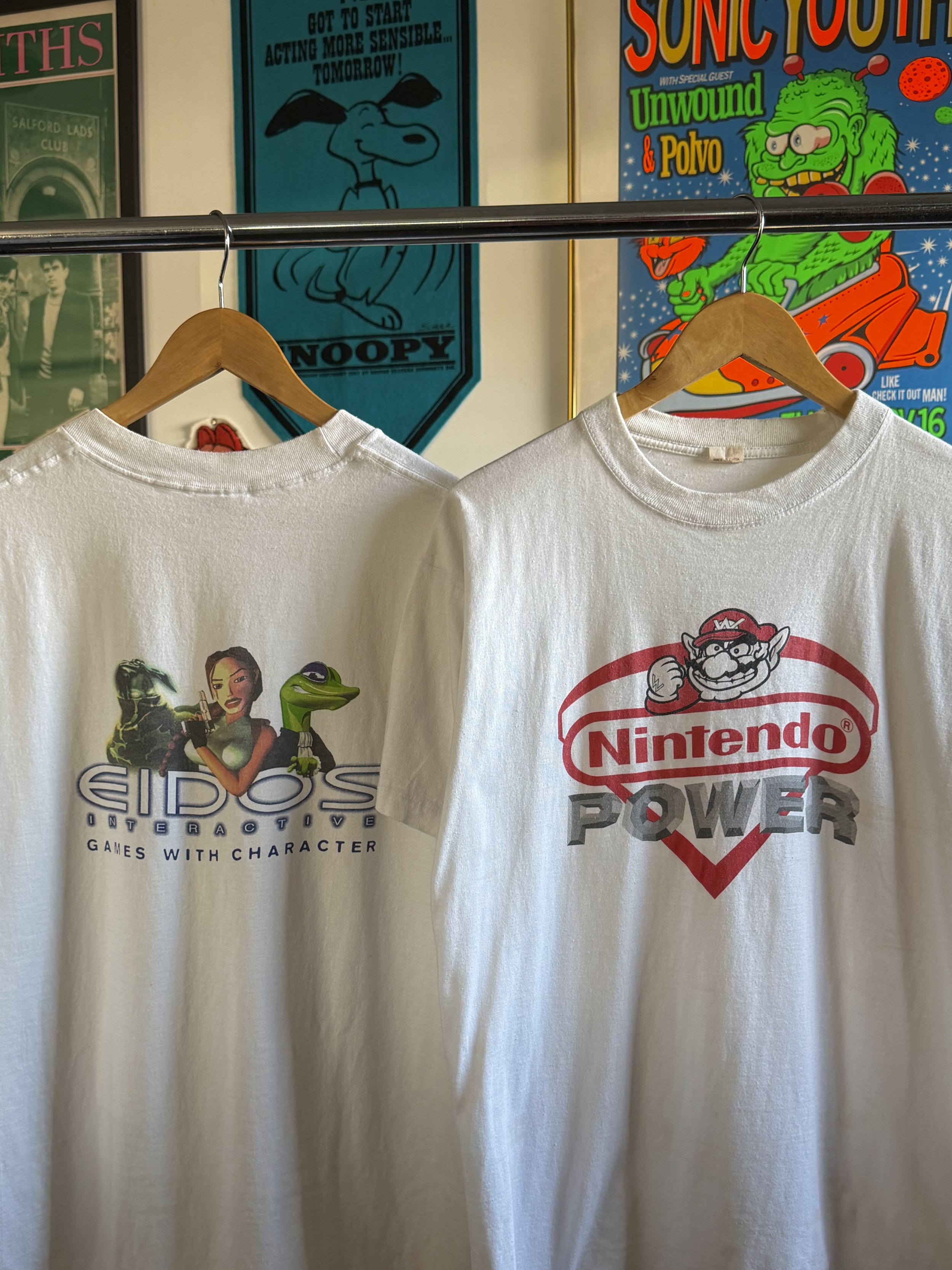Two white T-shirts hanging on a clothing rack. The shirt on the left features characters from the game EverQuest with the text "EQOS INTERACTIVE GAMES WITH CHARACTERS". The shirt on the right features Mario from Nintendo with the text "Nintendo Power