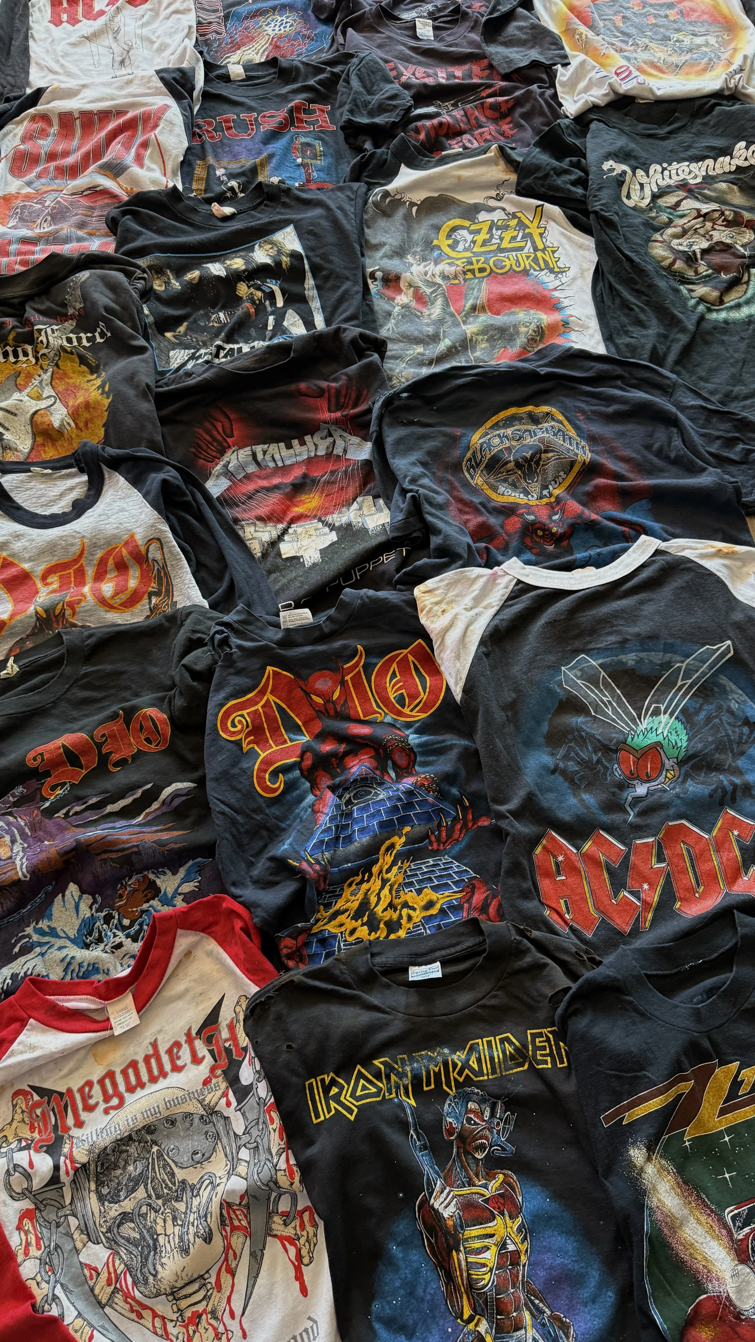 A collection of vintage rock band T-shirts, including designs from Metallica, AC/DC, Iron Maiden, Guns N' Roses, Queen, and others, arranged on a surface.