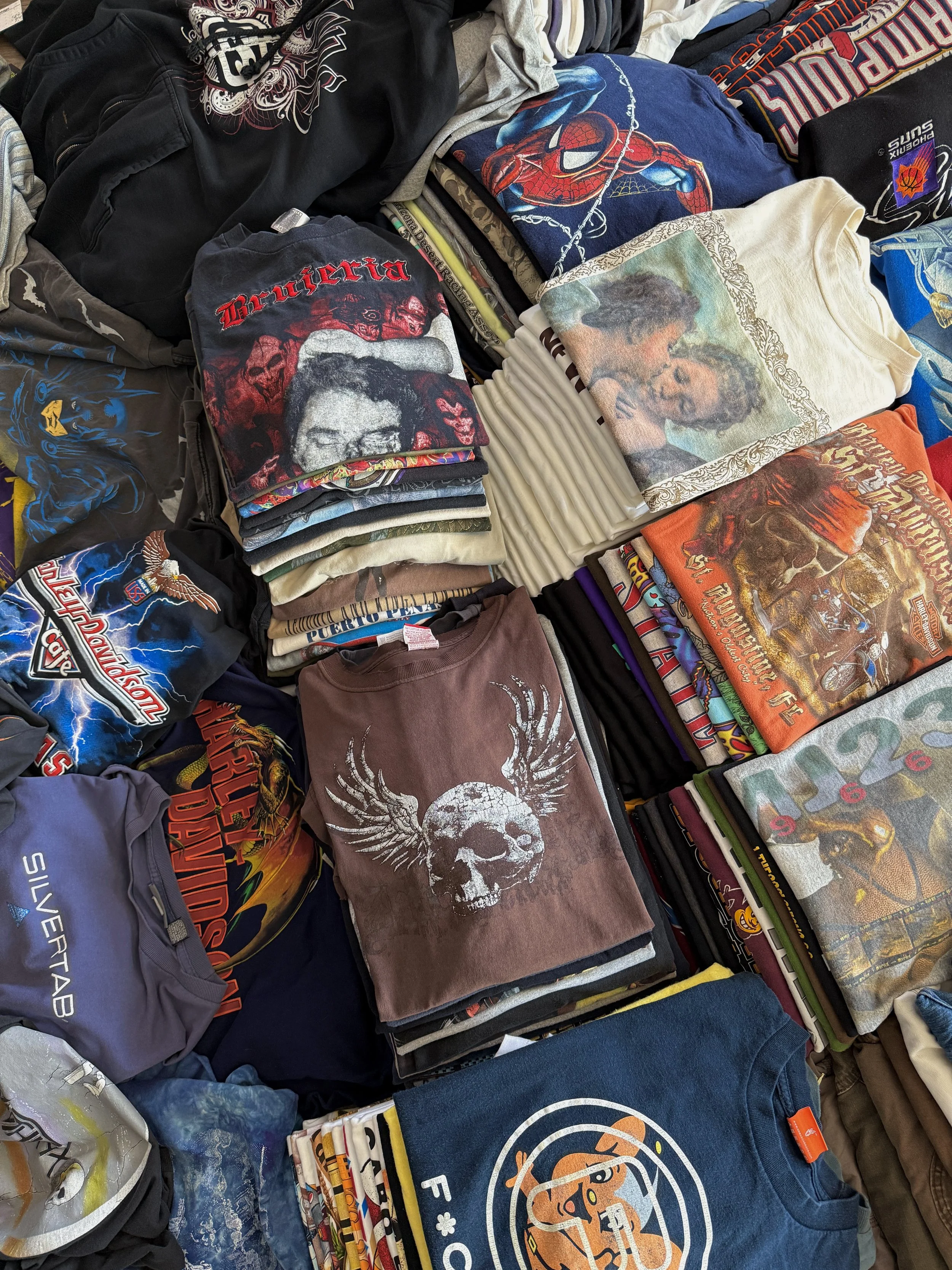 A collection of folded T-shirts with various graphic prints and designs, including horror movie promo, superhero, pop culture and music band tees, arranged on a flat surface.