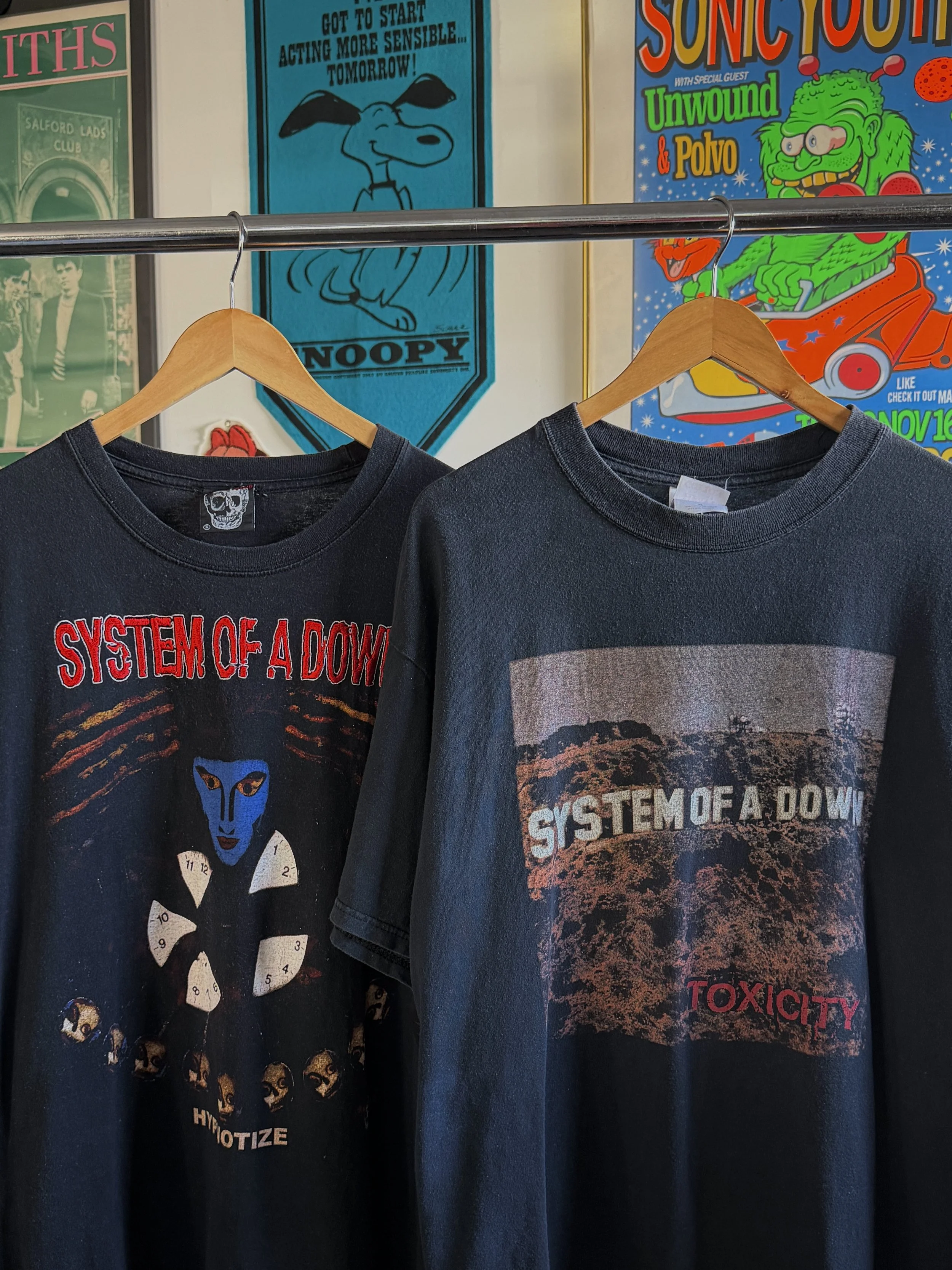 Two black System of a Down band t-shirts hanging on wooden hangers on a metal rod. The left shirt has a blue face with eight surrounding skulls and the words 'HYOTIZE' and 'SYSTEM OF A DOWN.' The right shirt shows an outdoor landscape with the words 