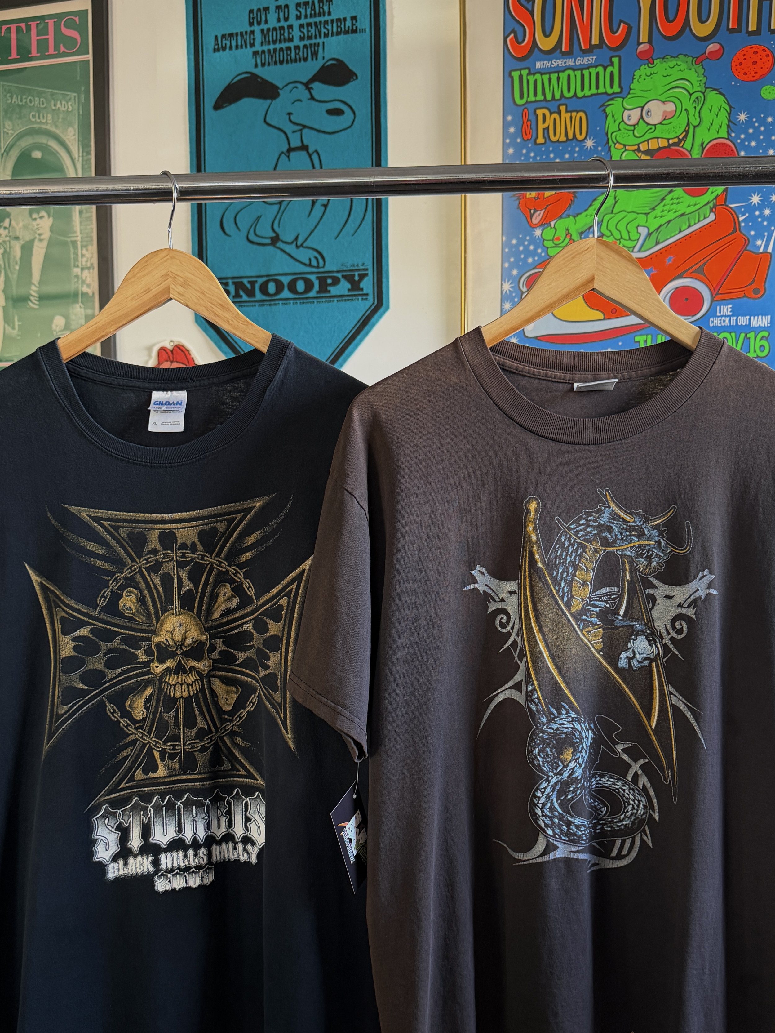 Two graphic t-shirts hanging on wooden hangers on a clothing rack in front of colorful posters, including one with Snoopy and another with Sonic Youth and other vibrant cartoon imagery.
