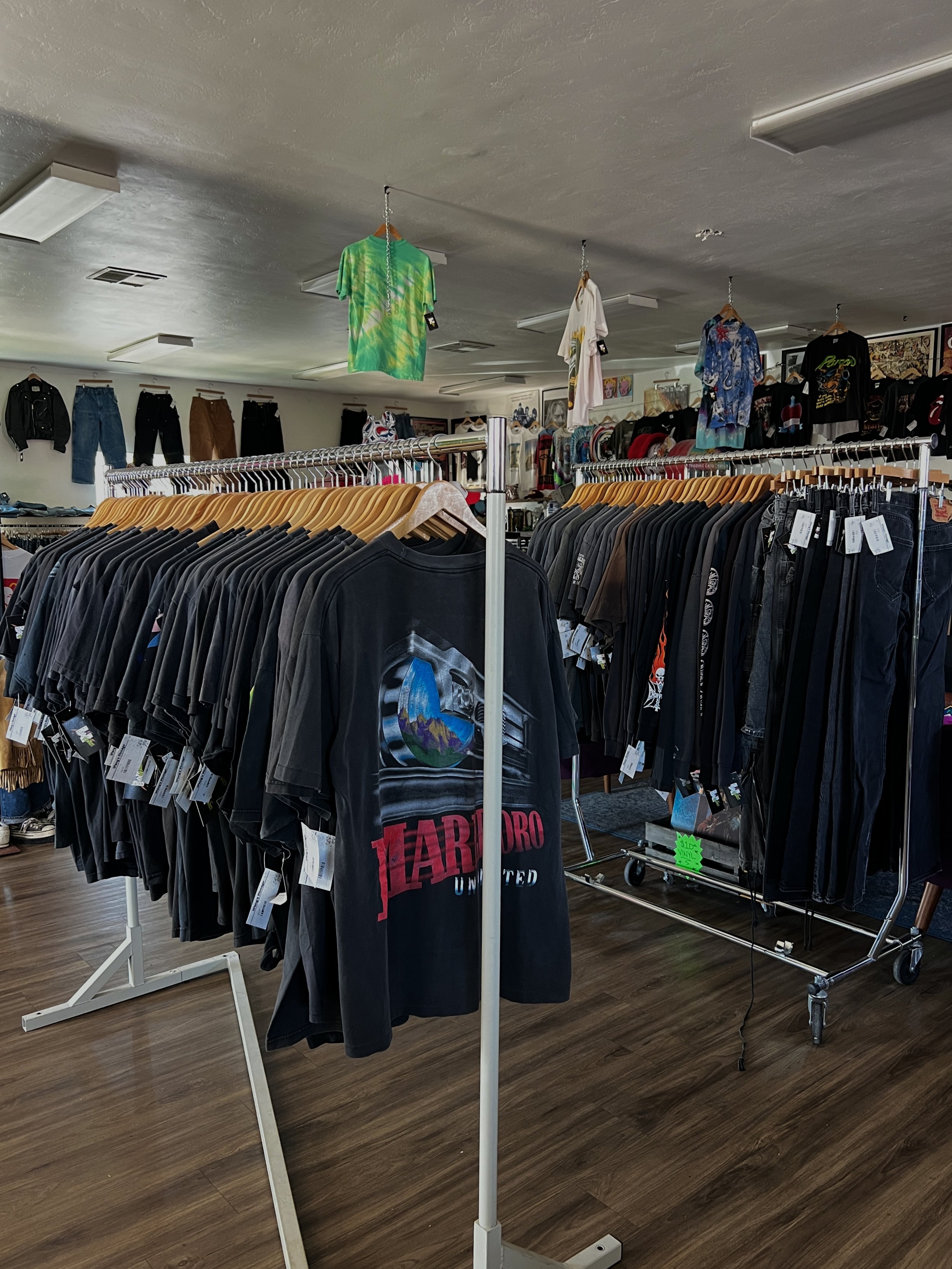 Inside a vintage clothing store with racks of mostly black and dark-colored T-shirts and pants, and some hanging tie-dye and graphic shirts, with vintage paintings and framed art on the walls in the background.