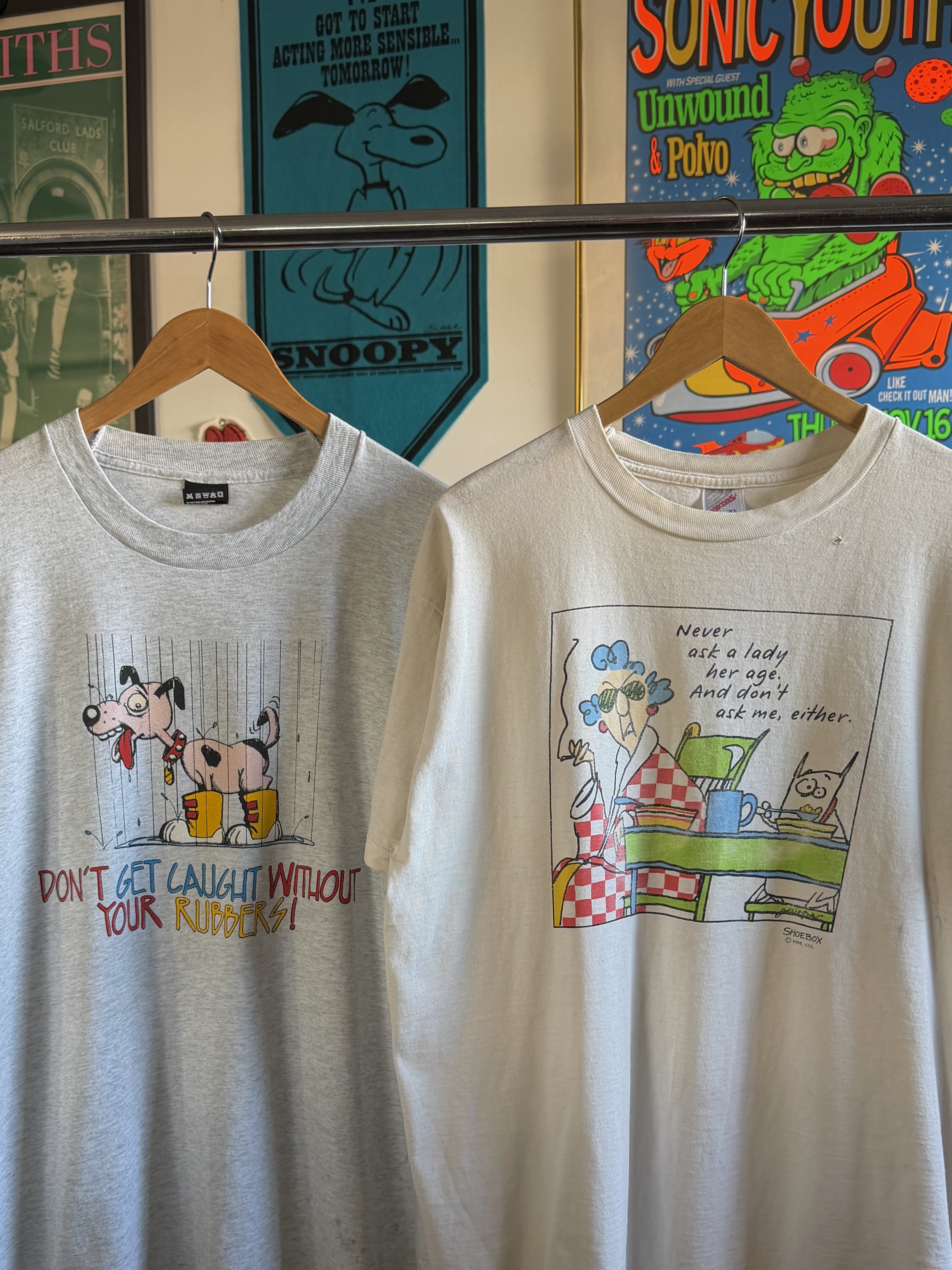 Two T-shirts hanging on a clothing rack with cartoon designs. The T-shirt on the left features a cartoon image of dogs and the phrase "DON'T GET CAUGHT WITHOUT YOUR RUBBERS!" The T-shirt on the right shows a cartoon woman with glasses and a cat, with