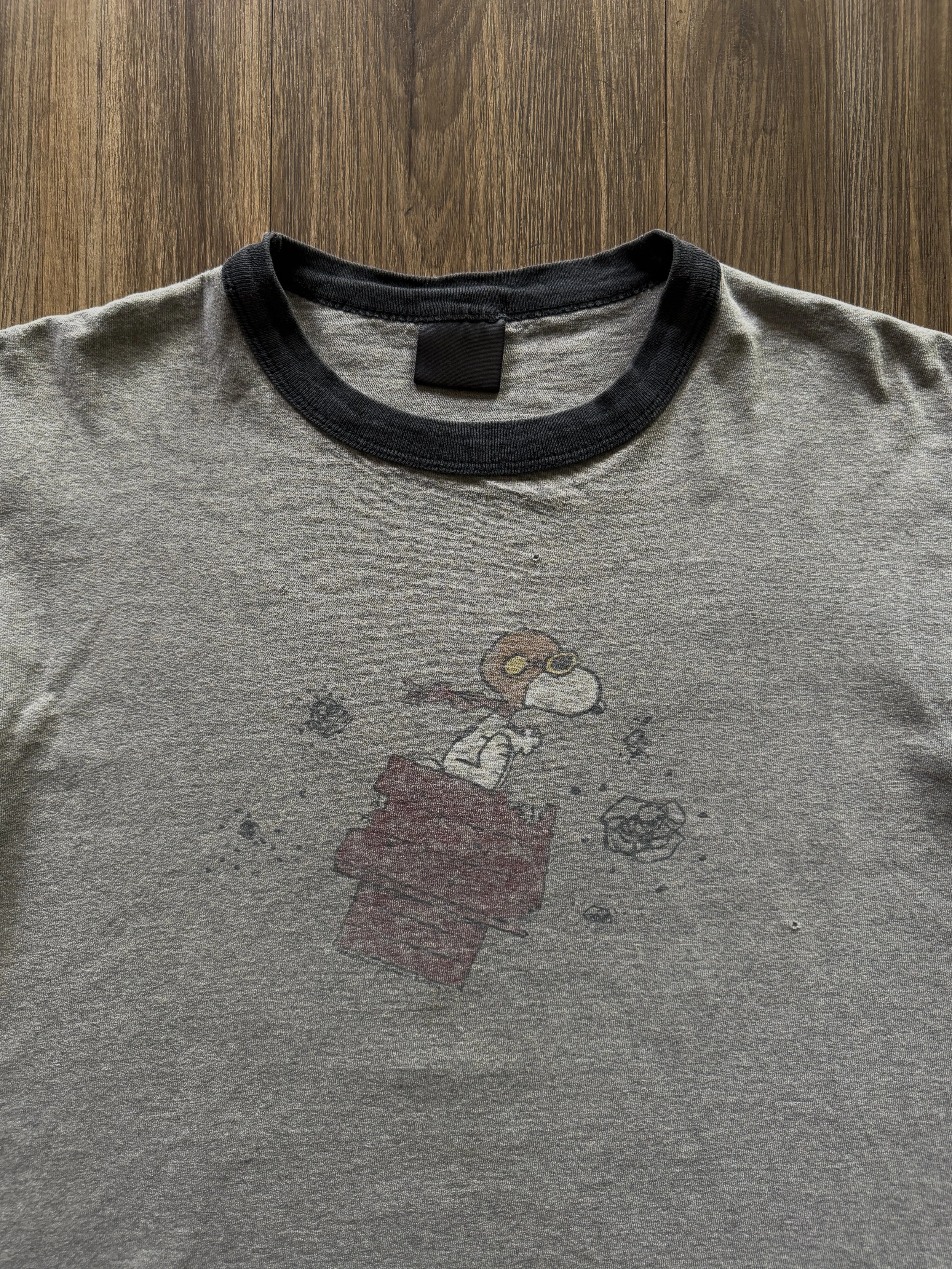 Gray T-shirt with black collar featuring a graphic of Snoopy from Peanuts wearing aviator goggles and scarf, sitting on top of a red doghouse, throwing rocks.