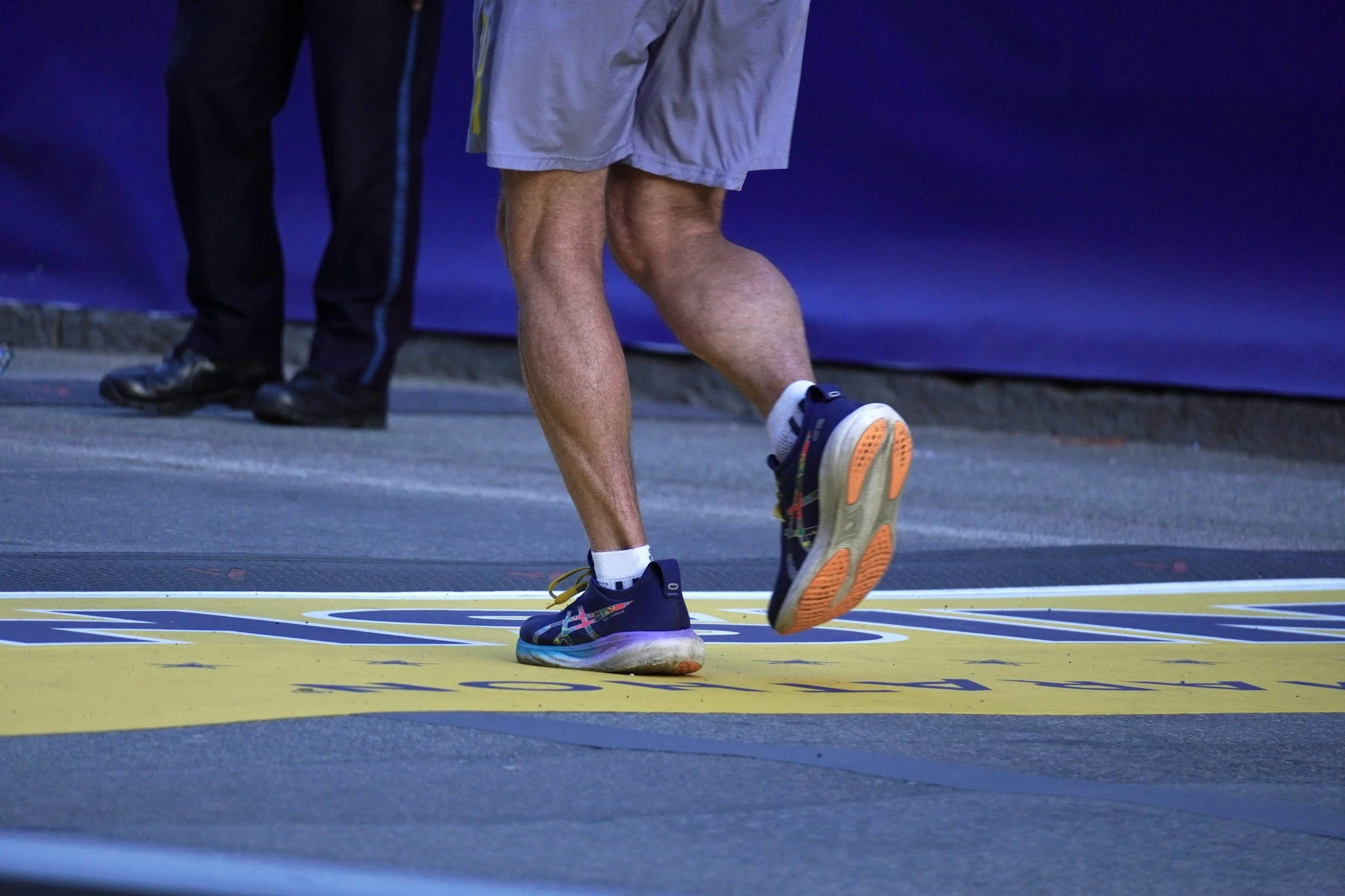Close-up of a runner's legs and feet crossing a finish line during a race, with a person standing in the background.