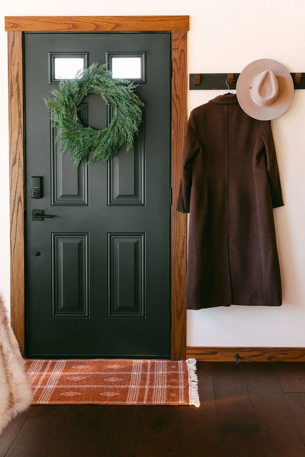 Dark green entry door with a wreath, a hat and coat on hooks and a runner rug