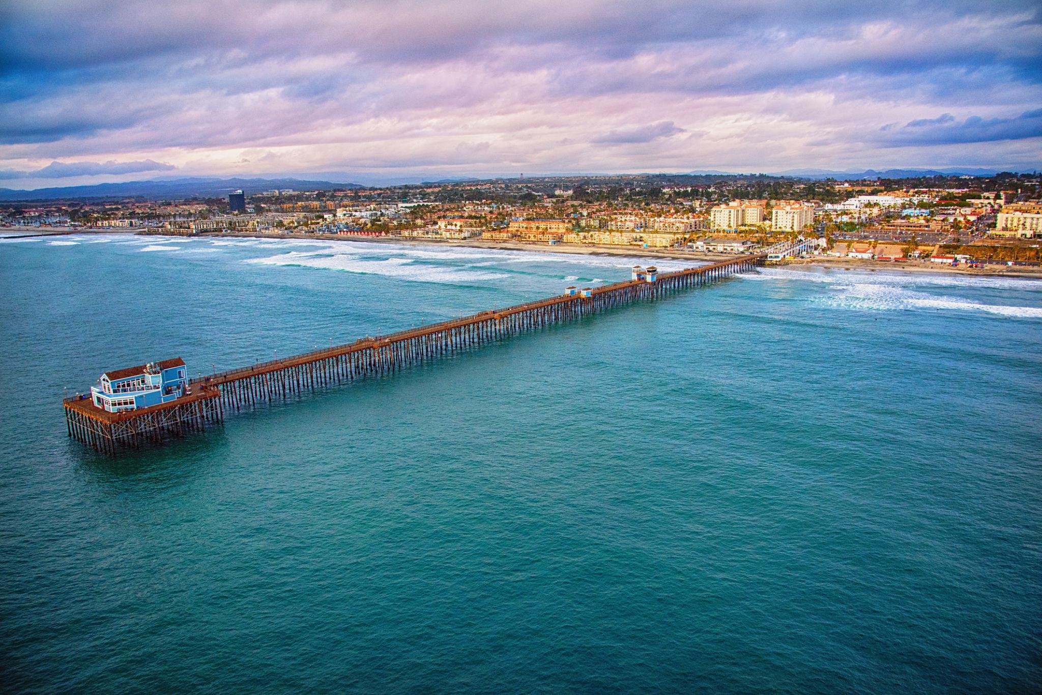 The Oceanside Pier reaching out over blue green water, seen from the air with the town and hills behind