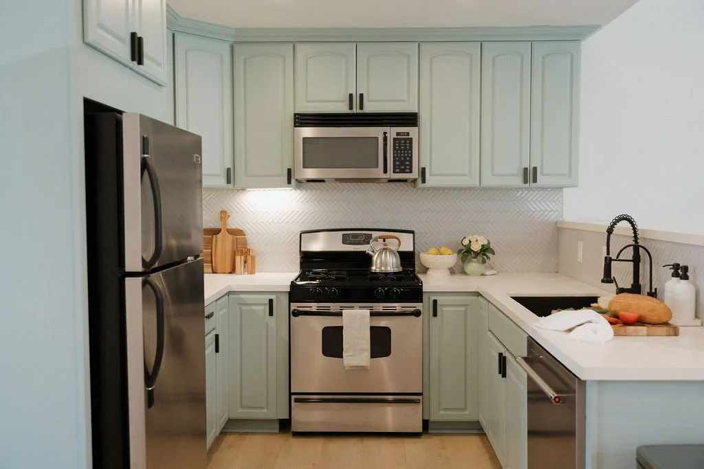 A modern kitchen with light blue cabinets, stainless steel refrigerator, microwave, oven, and a black farmhouse sink. Countertops have a bowl of lemons, a potted plant, and cutting boards with vegetables.