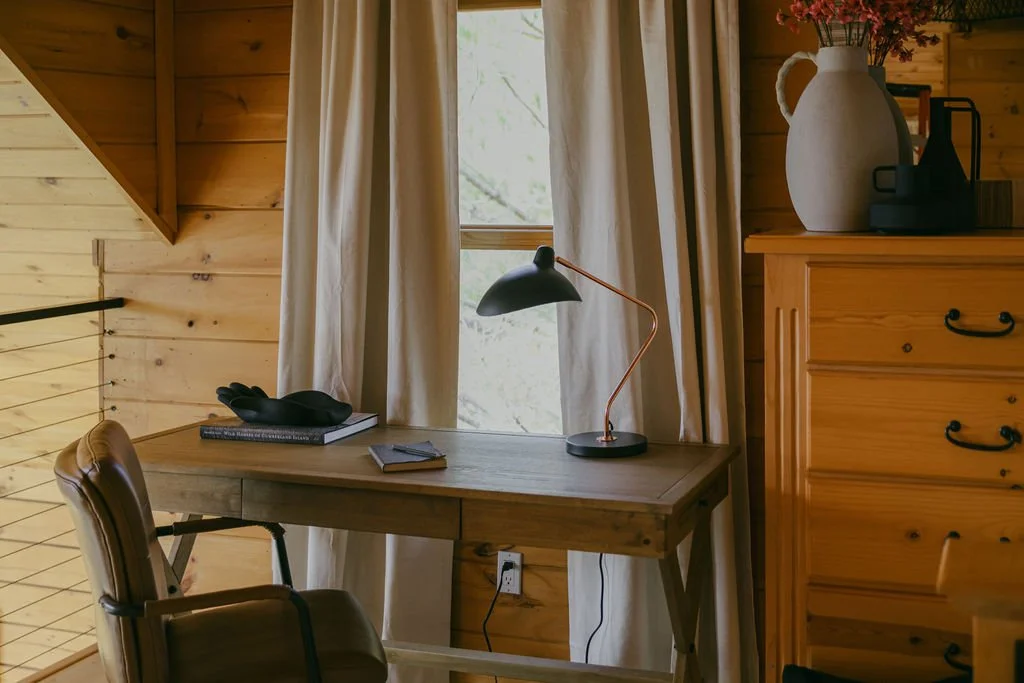 A wooden desk with a black and copper desk lamp, a pair of black gloves, a few books, and a small notepad. Behind the desk are cream curtains and a window revealing a green outdoor scene. To the right, there's a wooden dresser with a large white vase filled with pink flowers and some black office items.