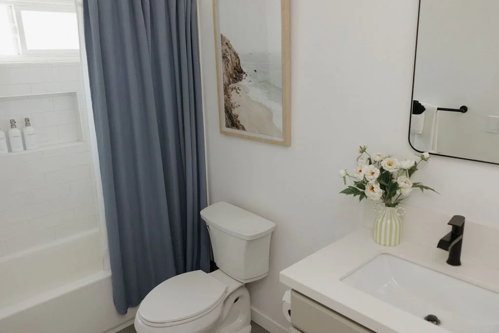 Small modern bathroom with white walls, a white toilet, a vanity with a white sink, a black faucet, a striped vase with white flowers, a mirror, a beach photo hanging on the wall, and a shower with a blue curtain.