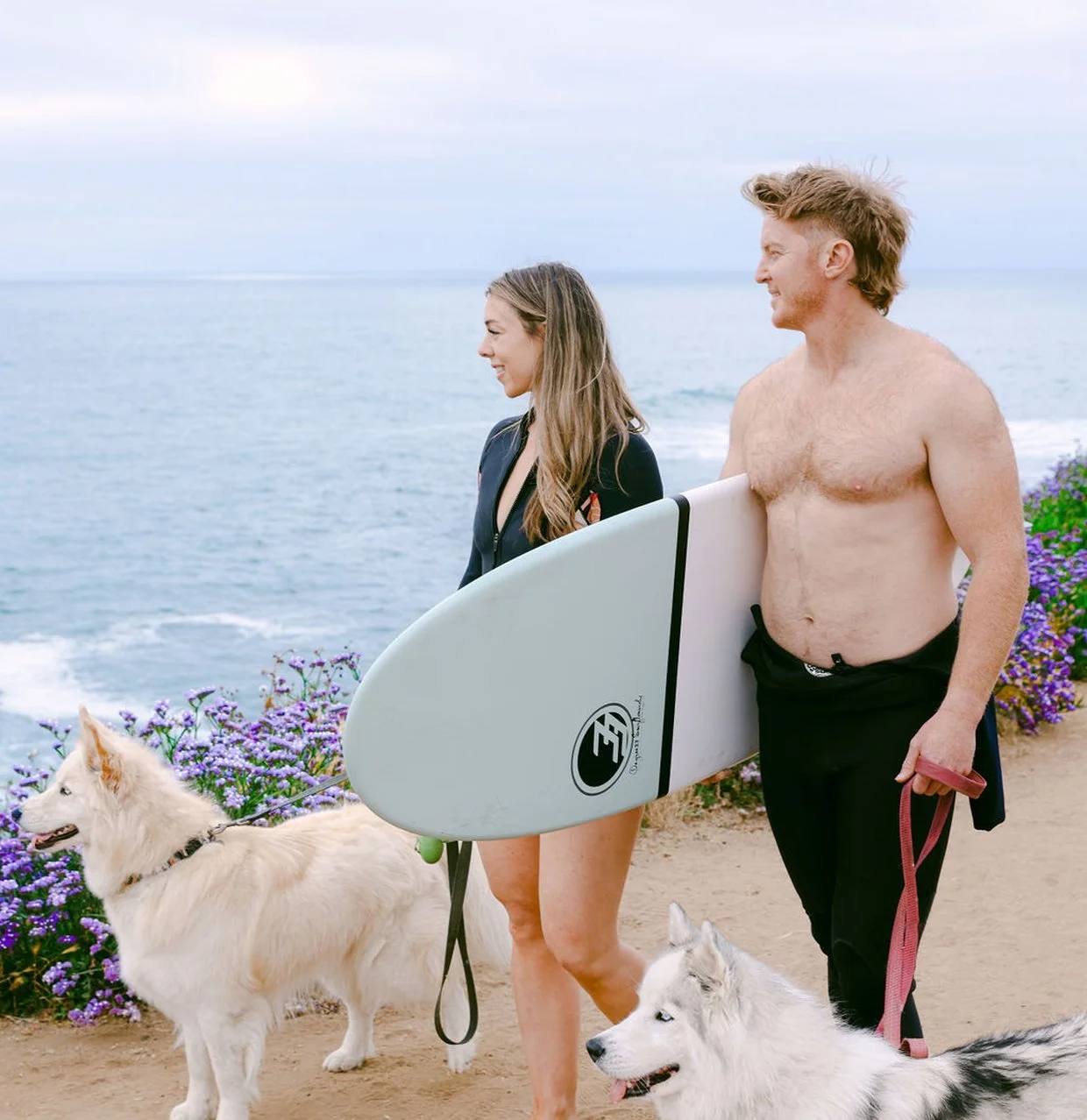 Aubrey and Brad walking up from the water with their two dogs and a surfboard, on the bluff above the ocean