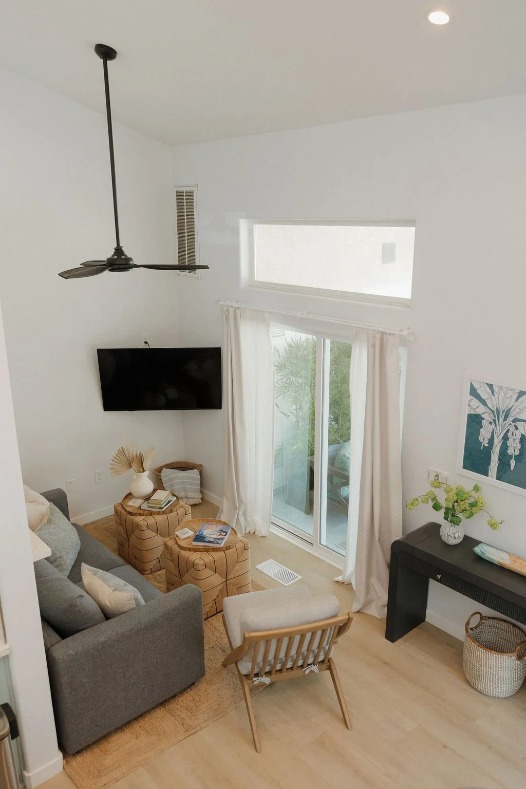 A living room with a gray sofa, two patterned ottomans, a wooden chair, a black console table with a vase of green flowers, a wall-mounted TV, a sliding glass door with white curtains, a rectangular window, and a ceiling fan.