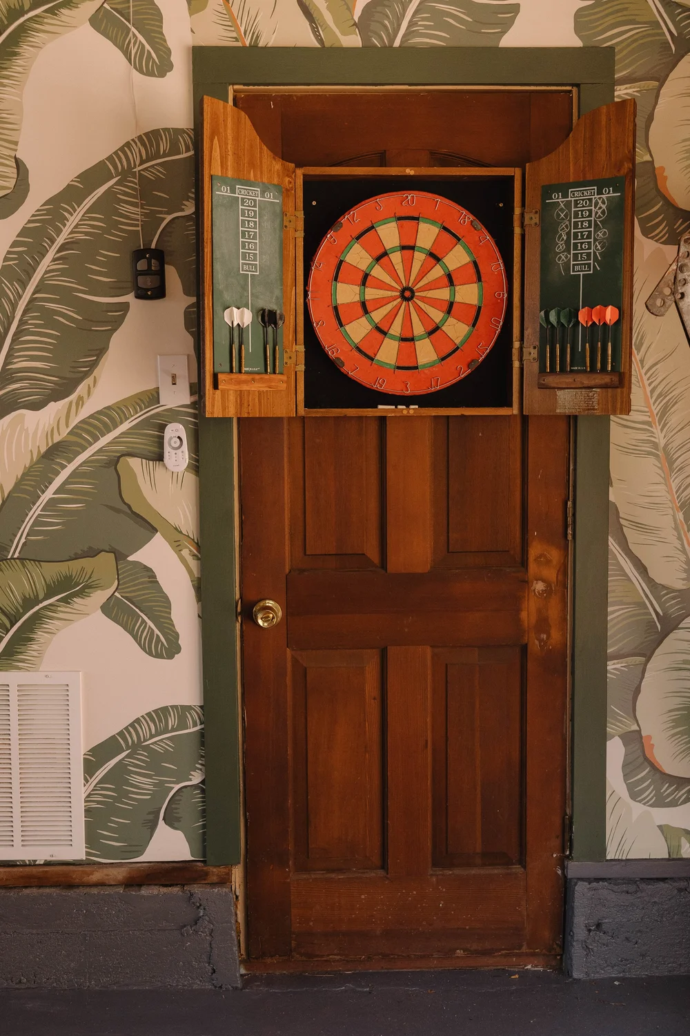 A dartboard on a wood door framed by palm leaf wallpaper in the game room at Palm Parkside