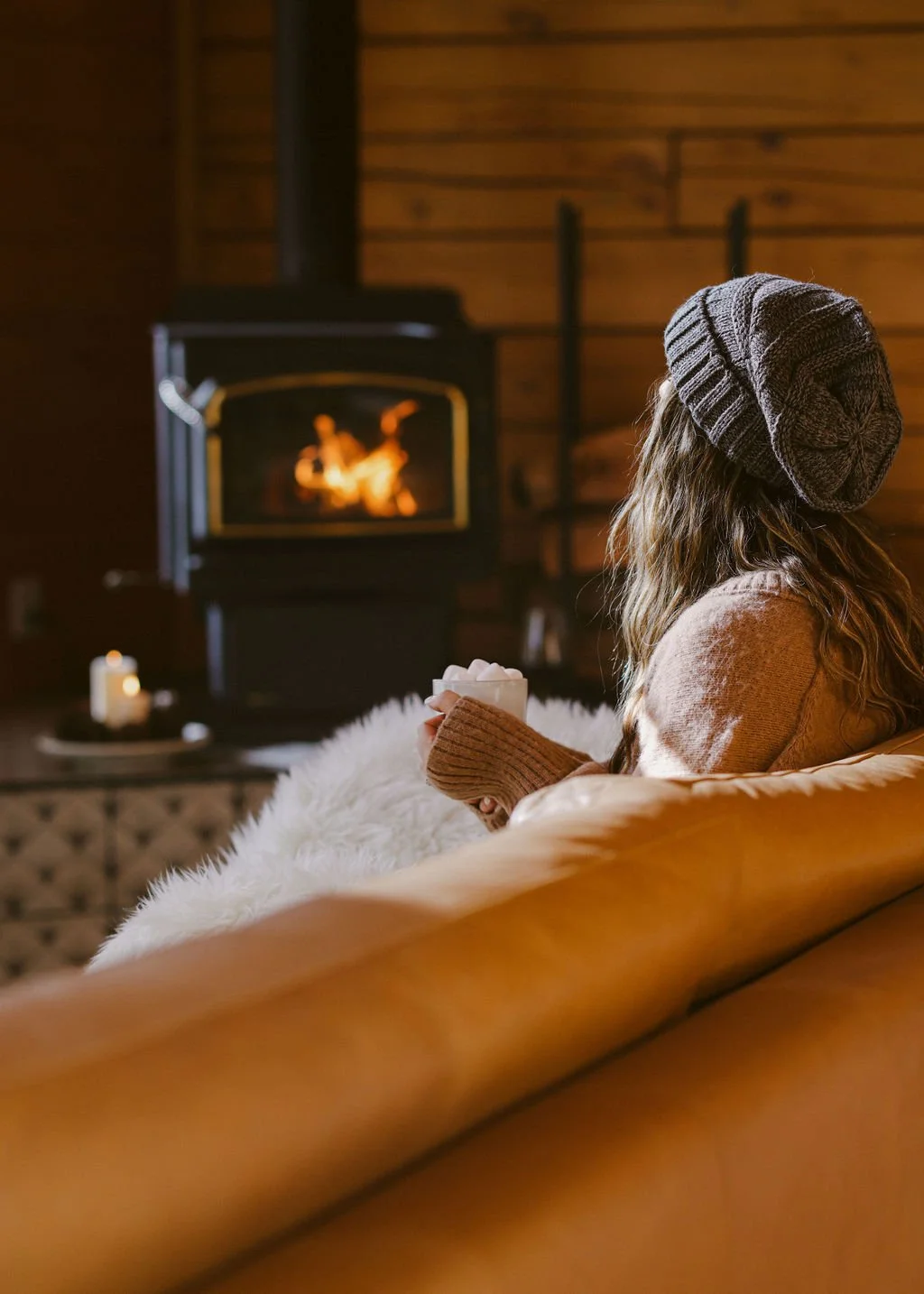 A woman with wavy hair wearing a gray knit beanie, sitting on a sofa with a white fuzzy blanket, holding a mug, and facing a lit fireplace in a cozy wood-paneled room.