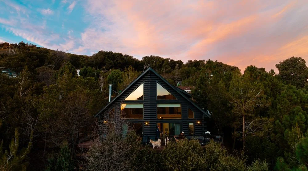 A cabin in the mountains near Julian lit from inside at dusk, with a dog on the deck and a pink and orange sky above the pines